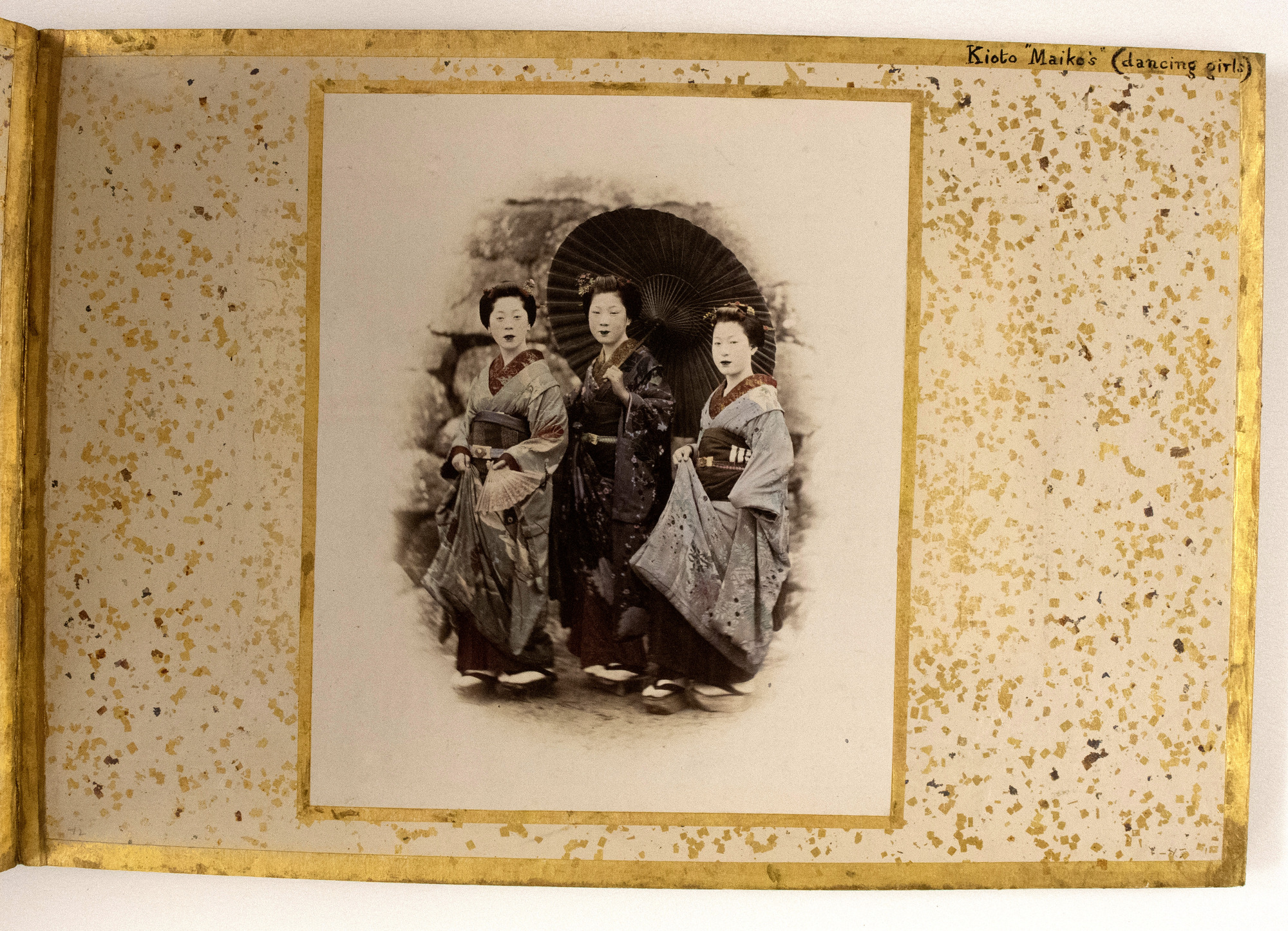 Sepia photograph colored with blue and red shades representing three standing Japanese women, the one in the center holding an open parasol. They are photographed outside against a stone wall, all three looking at the camera. The photograph is placed in the center of an album page decorated with a gold-leaf frame, colorful dots, and an inscription.