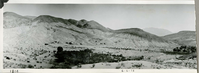 This is an historic black and white panoramic photograph from the Scotty's Castle Historic Photograph Collection, Death Valley National Park of building and landscaping development in desert landscape.  Extensive vegetation in mid-foreground that is the springs area. Extreme right is Scotty's Castle Stables expansion. In distance cabins and tents of Indian Camp. Black ink inscriptions along lower border. Right edge torn and missing.