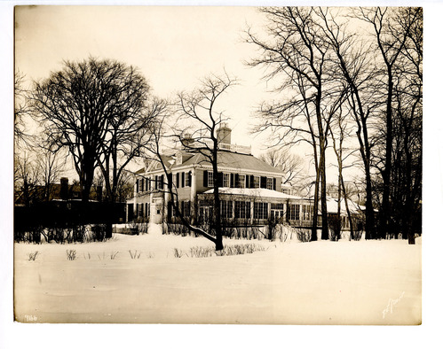 Black and white photograph of snowy landscape, featuring front and side of Georgian mansion.