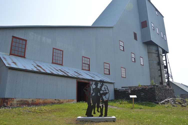 A metal sculpture of three men looks like they are exiting the door of the shaft-rock house that stands behind them. 