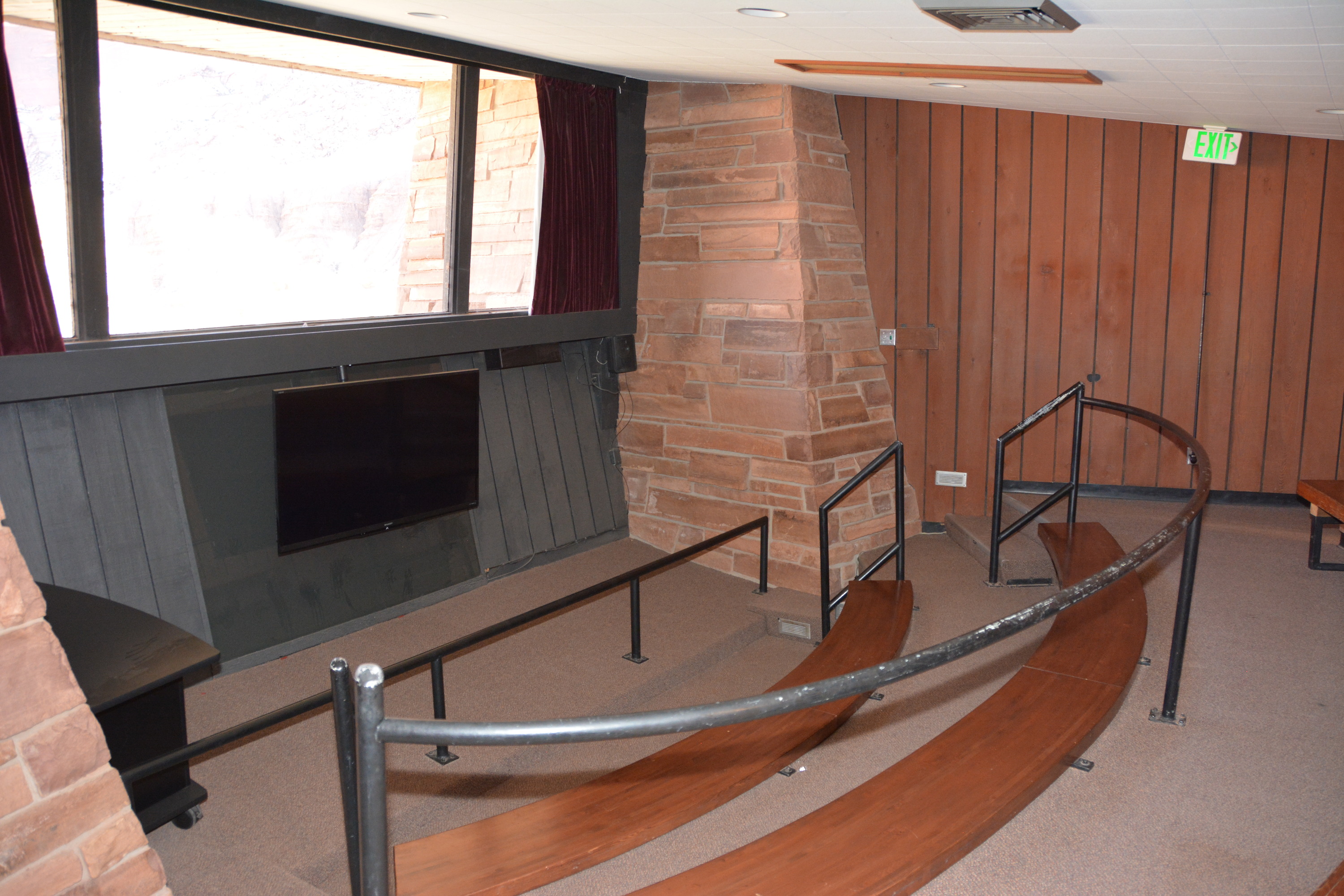 brown stone and wood theater with small screen and two rows of curved wooden benches, with carpeted floor, and a large window above the small screen. 