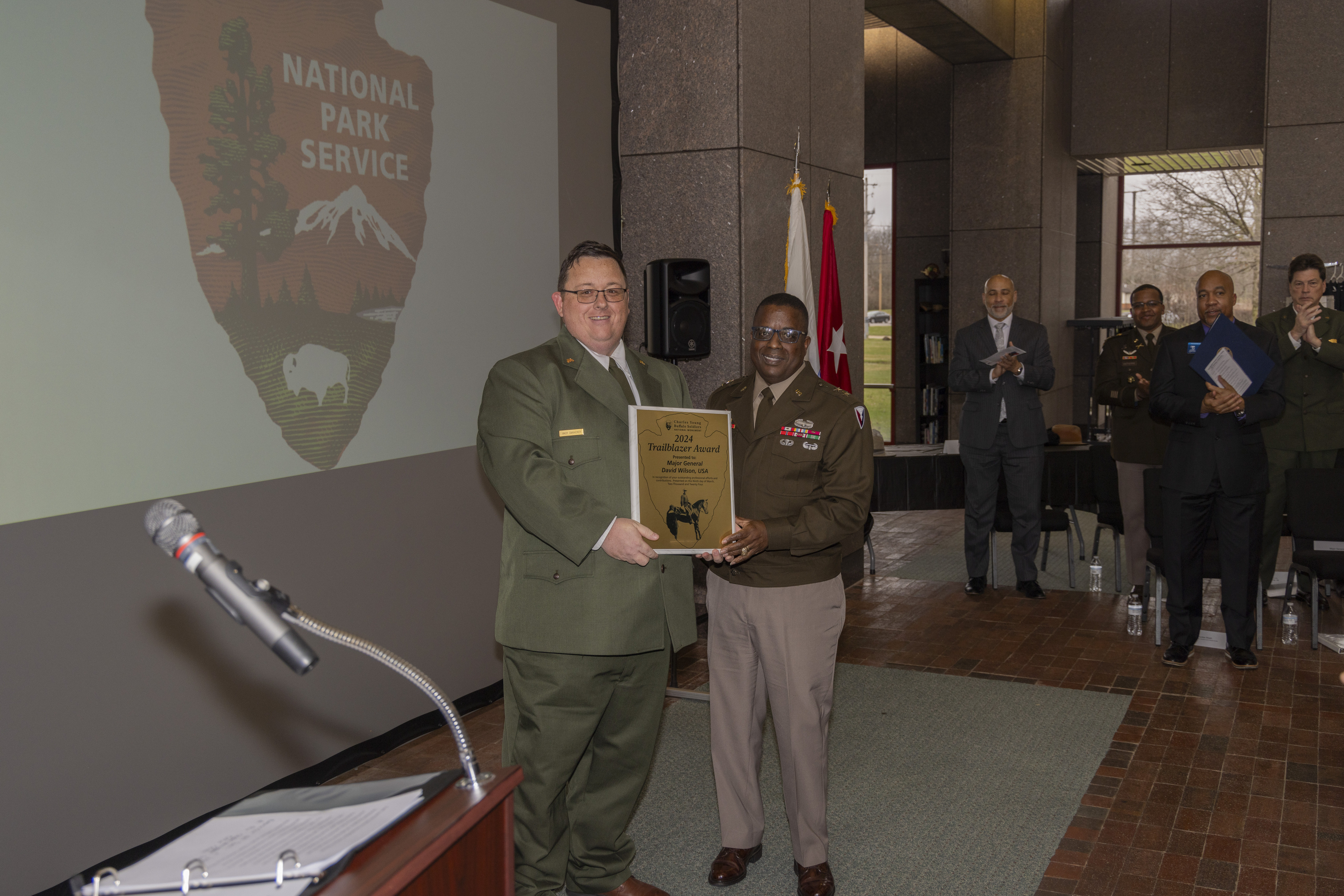A man in a dark brown military uniform suit on the right and a man in a green ranger suit on the left stand next to one another while both hold onto a brass-colored plaque with an image on the bottom of a man on a horse and the outline of an arrowhead around him. 
