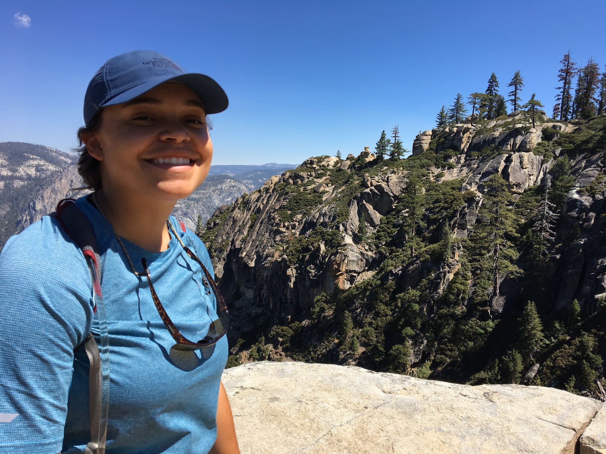 A woman in blue hat and shirt poses on the left side of the photo with overlooks of Yosemite National Park in the background.
