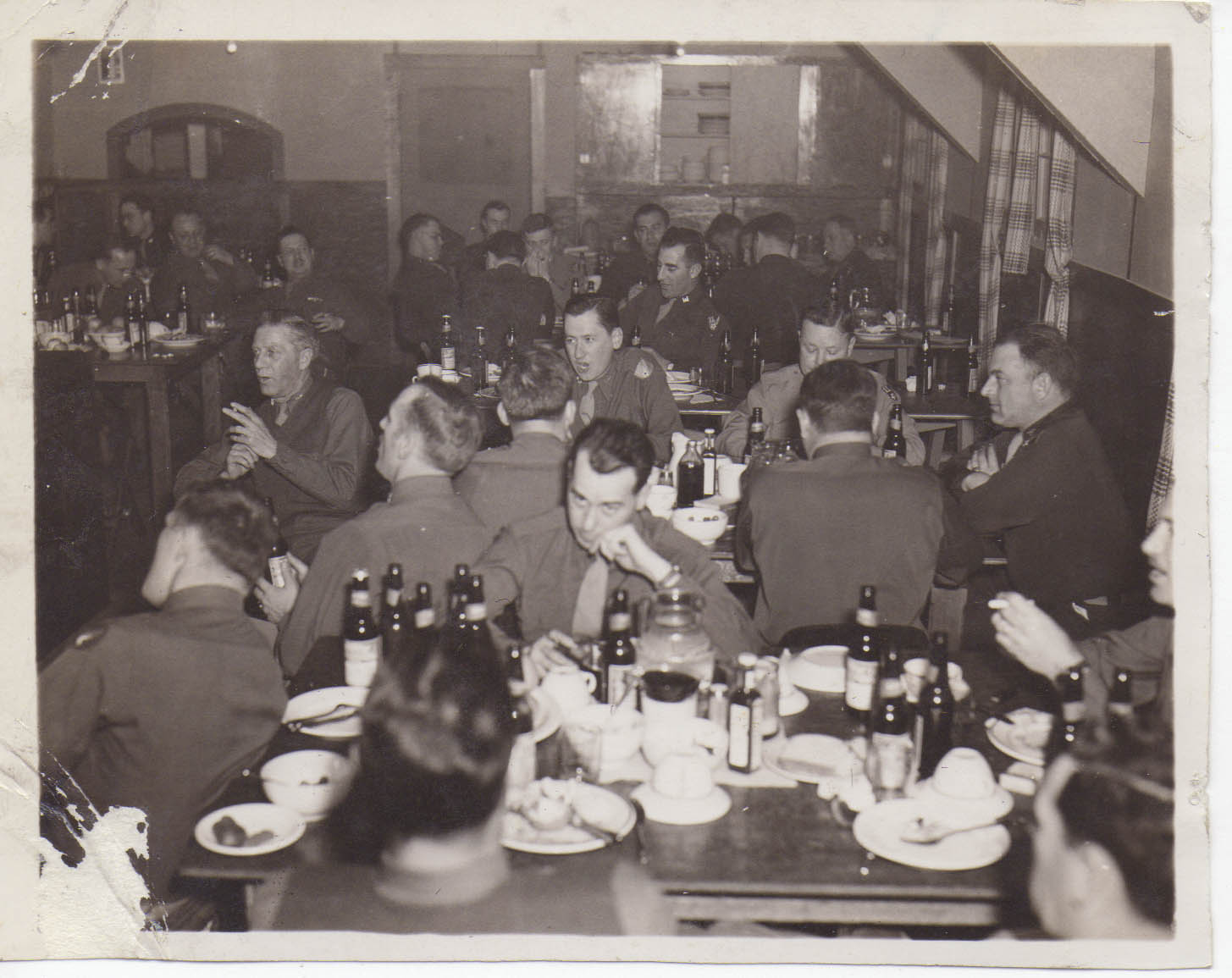 Uniformed men gathered around tables in a room.  Each table has many beer bottles on it.