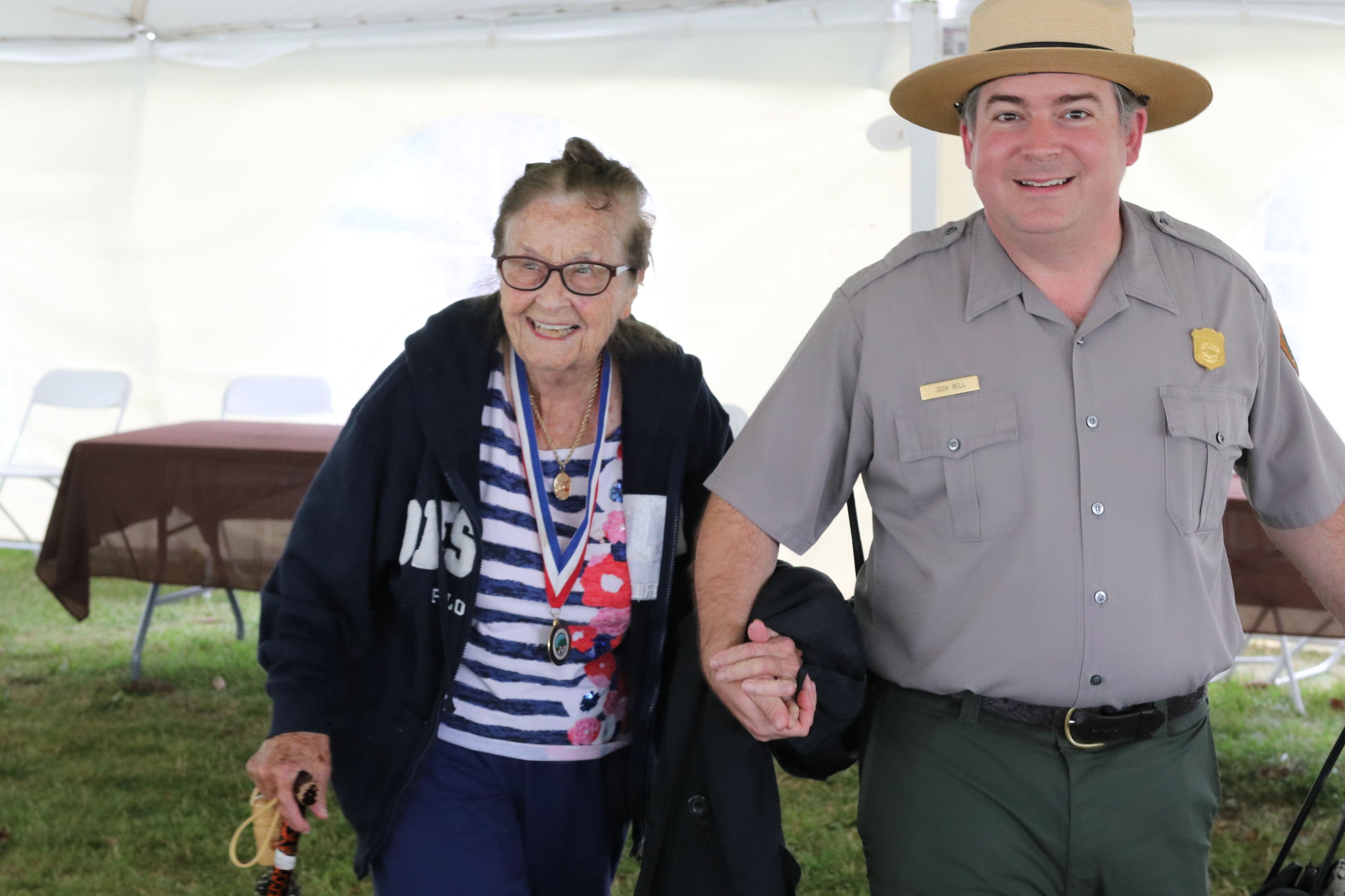 A park ranger in uniform holds hands with a WWII veteran while walking together. 