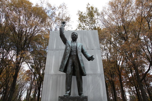 A tall bronze statue of  Theodore Roosevelt stands in front of several trees in the fall time.