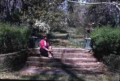 Four brick steps with matching urns on both sides of the top step.  Steps lead to path surrounded by shrubs and flowers.  A lady is sitting on the steps.
