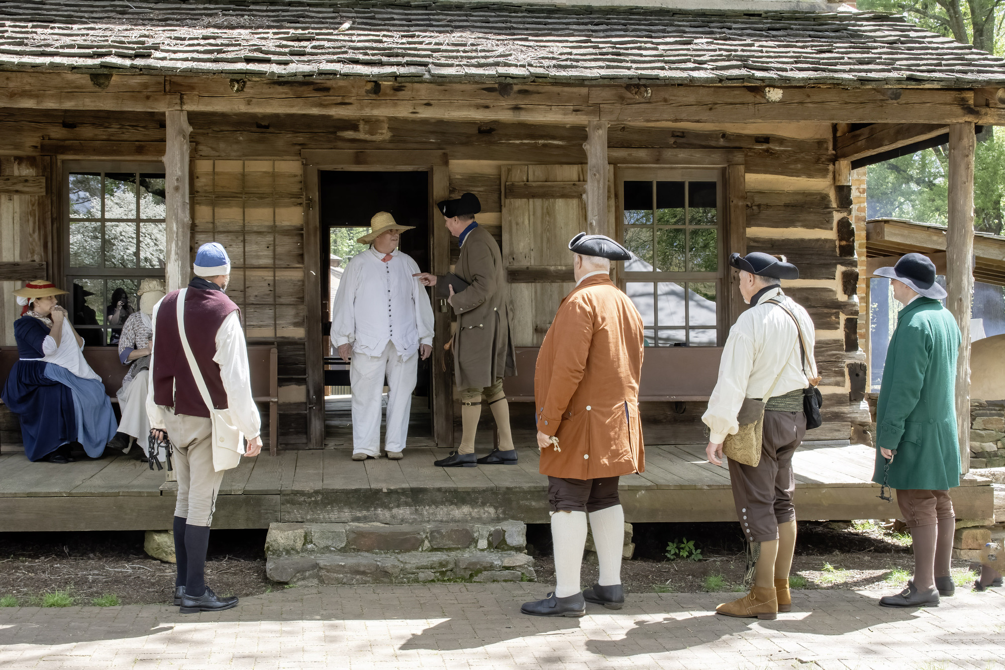 8 people in 18th century living history clothing stand and sit in front of a cabin. 