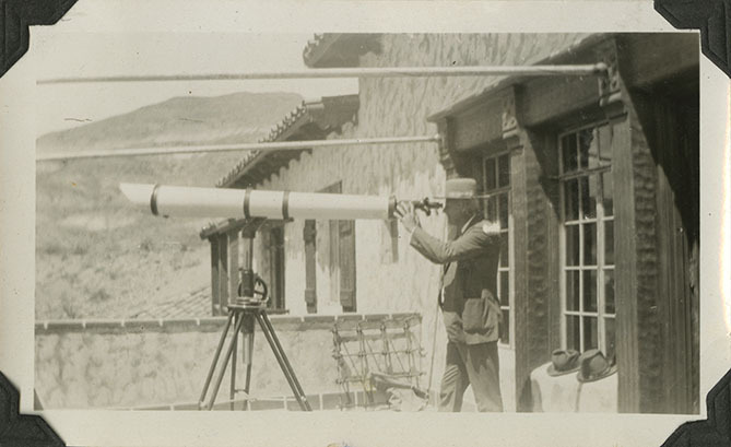 This is an historic black and white photograph from the Scotty's Castle Historic Photograph Collection, Death Valley National Park of Mat Roy Thompson with telescope on Scotty's Castle Main House Gallery Porch, looking west. Circa 1930.