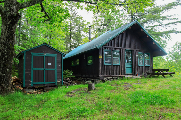 Large log cabin with a picnic table in front. A woodshed is between a tree and the cabin.