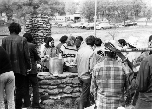 A large group of people stand outdoors facing away from the camera, waiting in line for food being served at a stone hearth