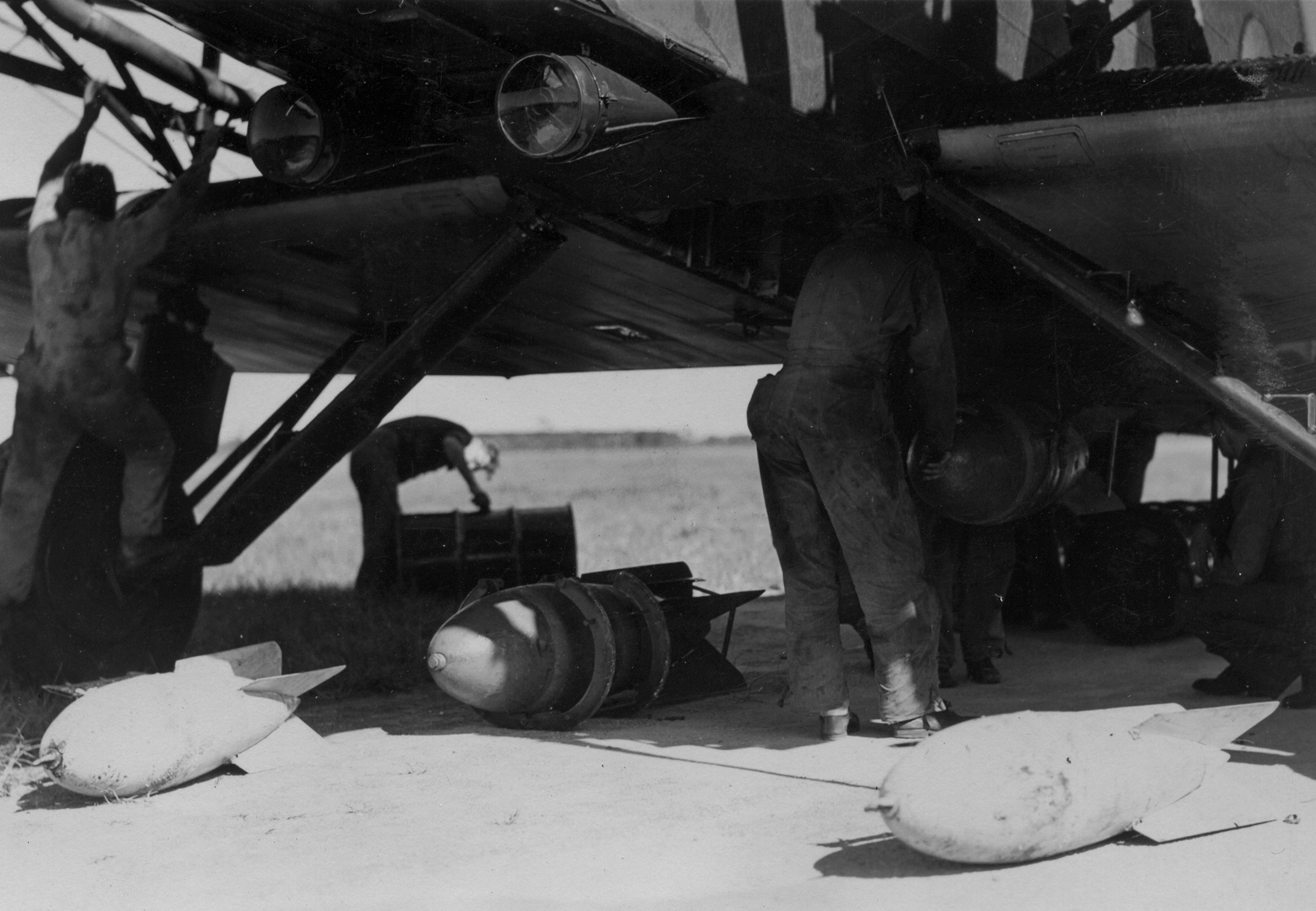 Black and white photo of bombs on the ground at the foot of a plane with three soldiers tending to the plane