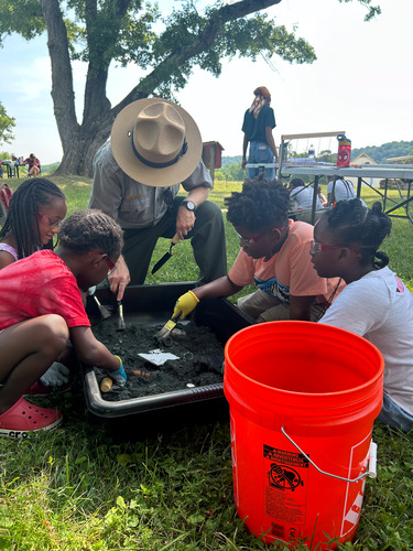 Park ranger with four smiling children. All of them are digging in the dirt using brushing and archaeological tools.