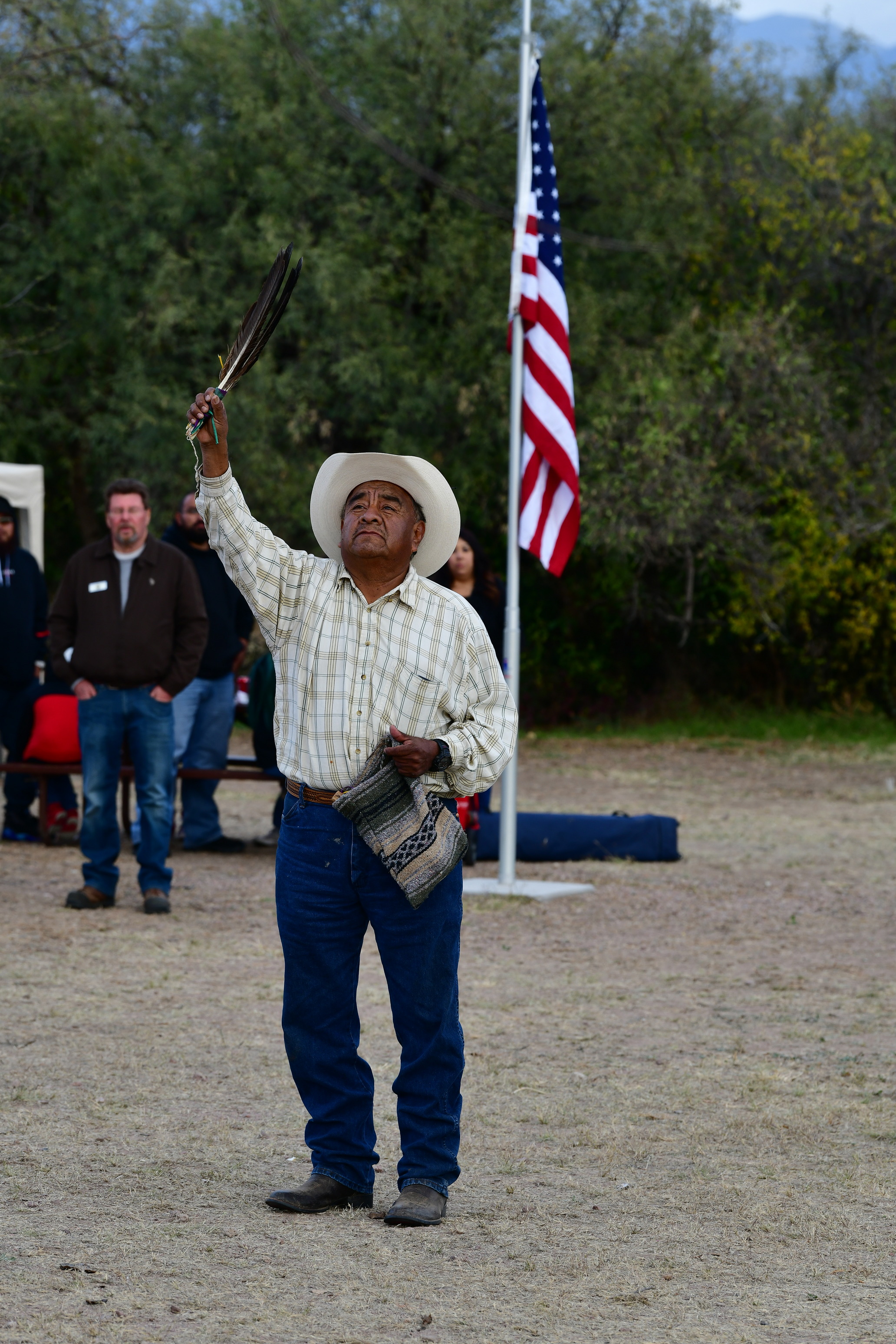 O'odham elder raises eagle feathers in blessing, American flag visible in background