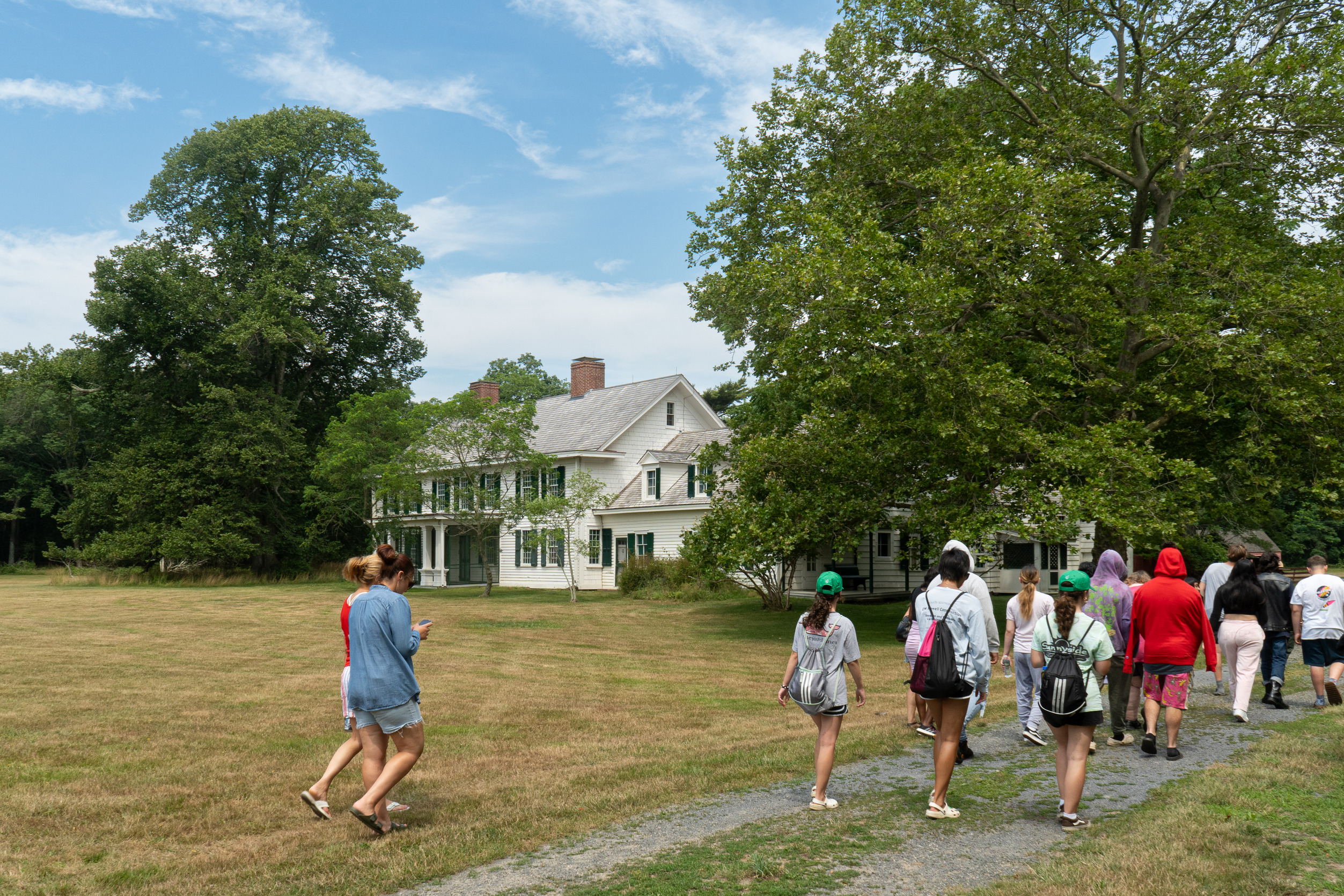 A group of students walk past a white house with green shutters on a grassy field.