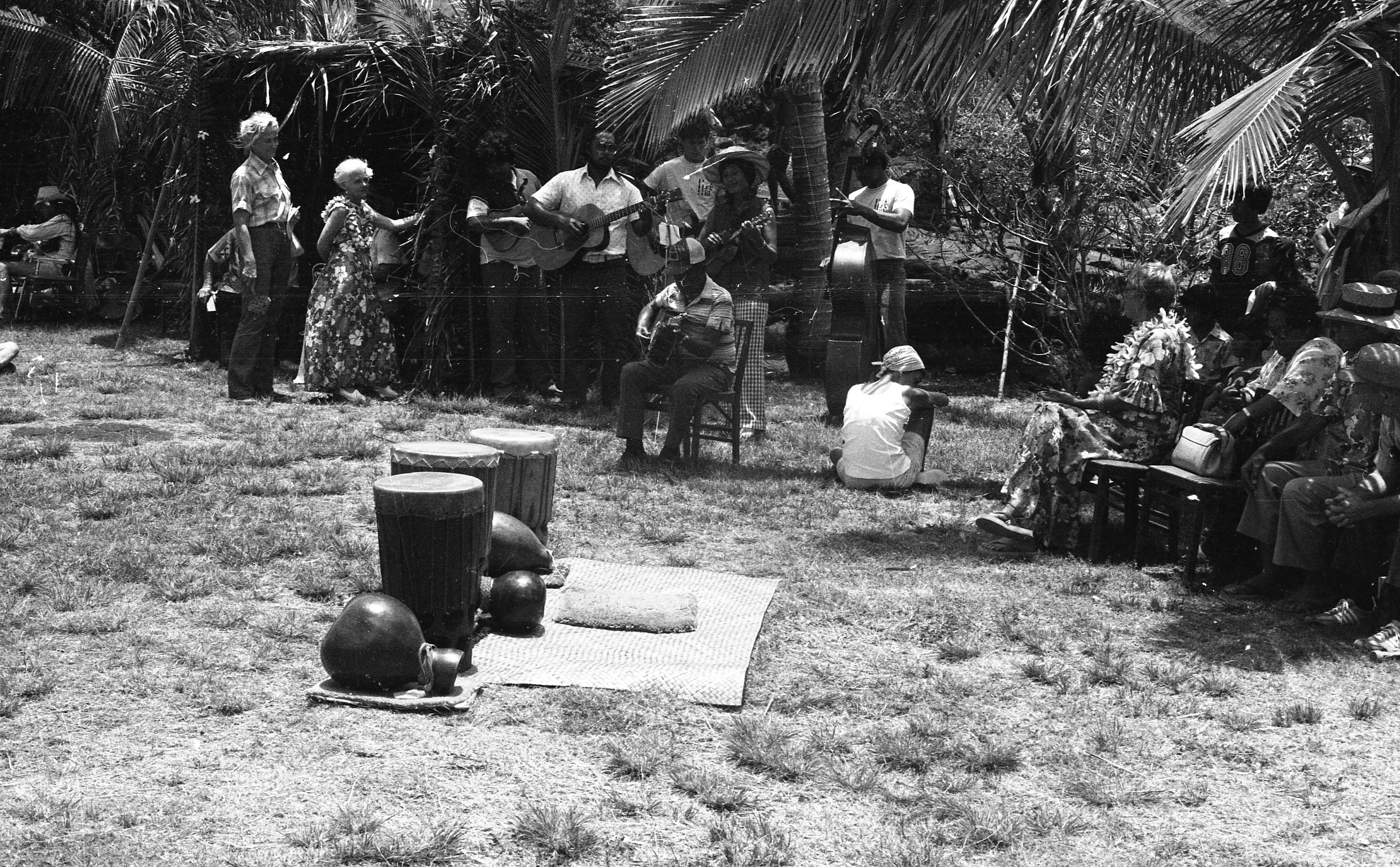 A black and white wide image of an event. In the center of the image there are three drums and three gourds lined up on a woven mat. Behind the instruments there is a cushion on the woven mat as well. On the right side of the image there is a group of people sitting in chairs. They are looking away from the camera towards a band located in the upper center of the image. All six members of the band are playing instruments. One man standing towards the seated audience is playing a bass while the others are playing guitars. On the left side of the band there is an older man and woman watching the band. The woman is wearing a dress with a floral design and a lei. She is holding onto a palm tree with her left hand. To the right of her is a man. He is wearing a short sleeve collared shirt and pants. In the background there is dense vegetation.