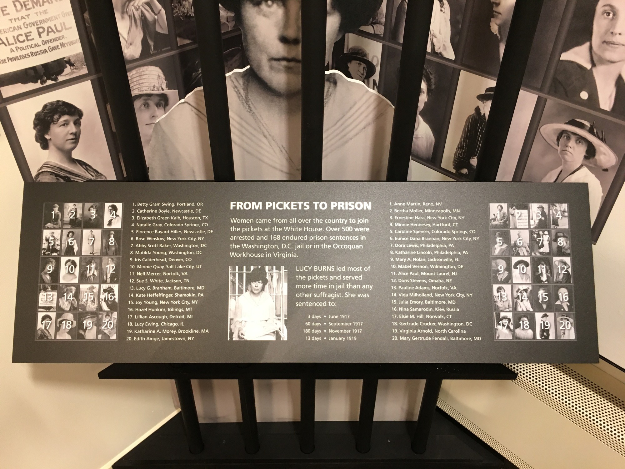 A zoomed in photo of the exhibit panel identifying the women's photos. A larger Lucy Burns cut out of her upper body sits behind prison bars. Behind her are photos of other women imprisoned. The sign explaining who is who is in front of the bars. 