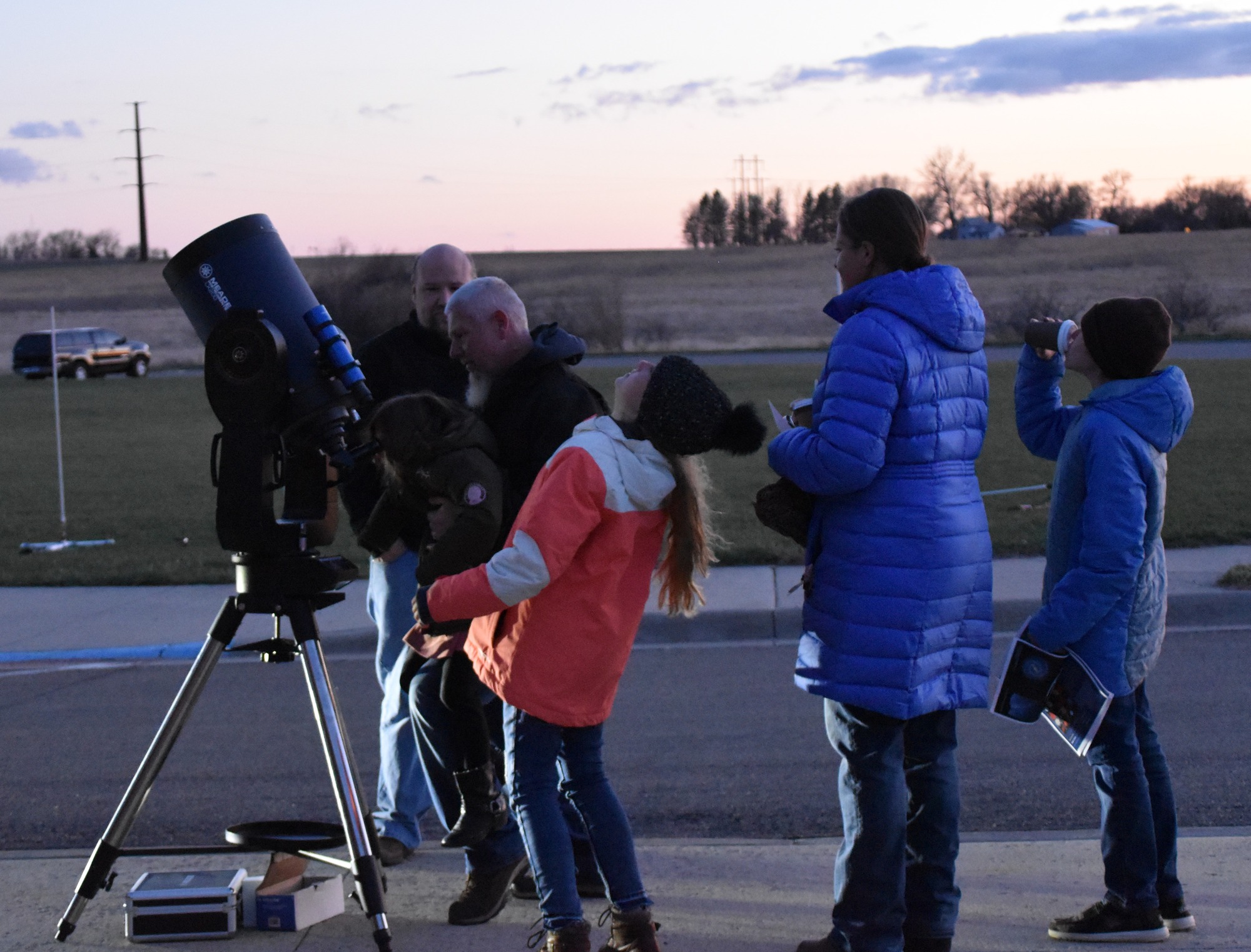 A man holds a child up to a telescope in front of a woman and two other children while a different man stands to the side. 