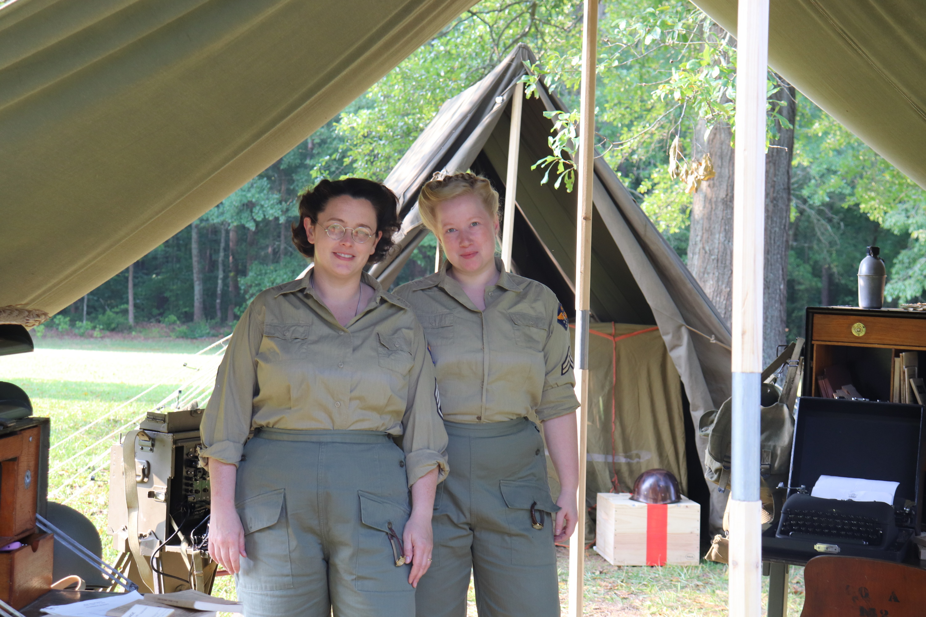 Two women dressed as WACs pose inside of a canvas tent with various period items such as typewriters and radios that they used to communicate with other military groups.
