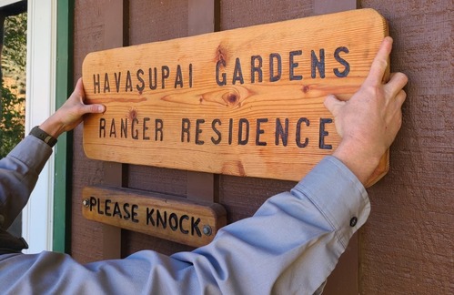 The arms of a ranger are seen mounting a sign outside of the Ranger Station which reads, "Havasupai Gardens Ranger Residence"