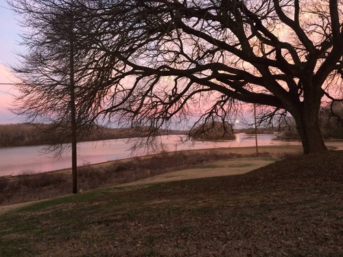View from the Dover Hotel (Surrender House) through bare tree branches toward the west as the sun is setting below a bank of clouds. The sky is colorful and is reflected off the surface of the water. Several forested islands are within the river channel. 