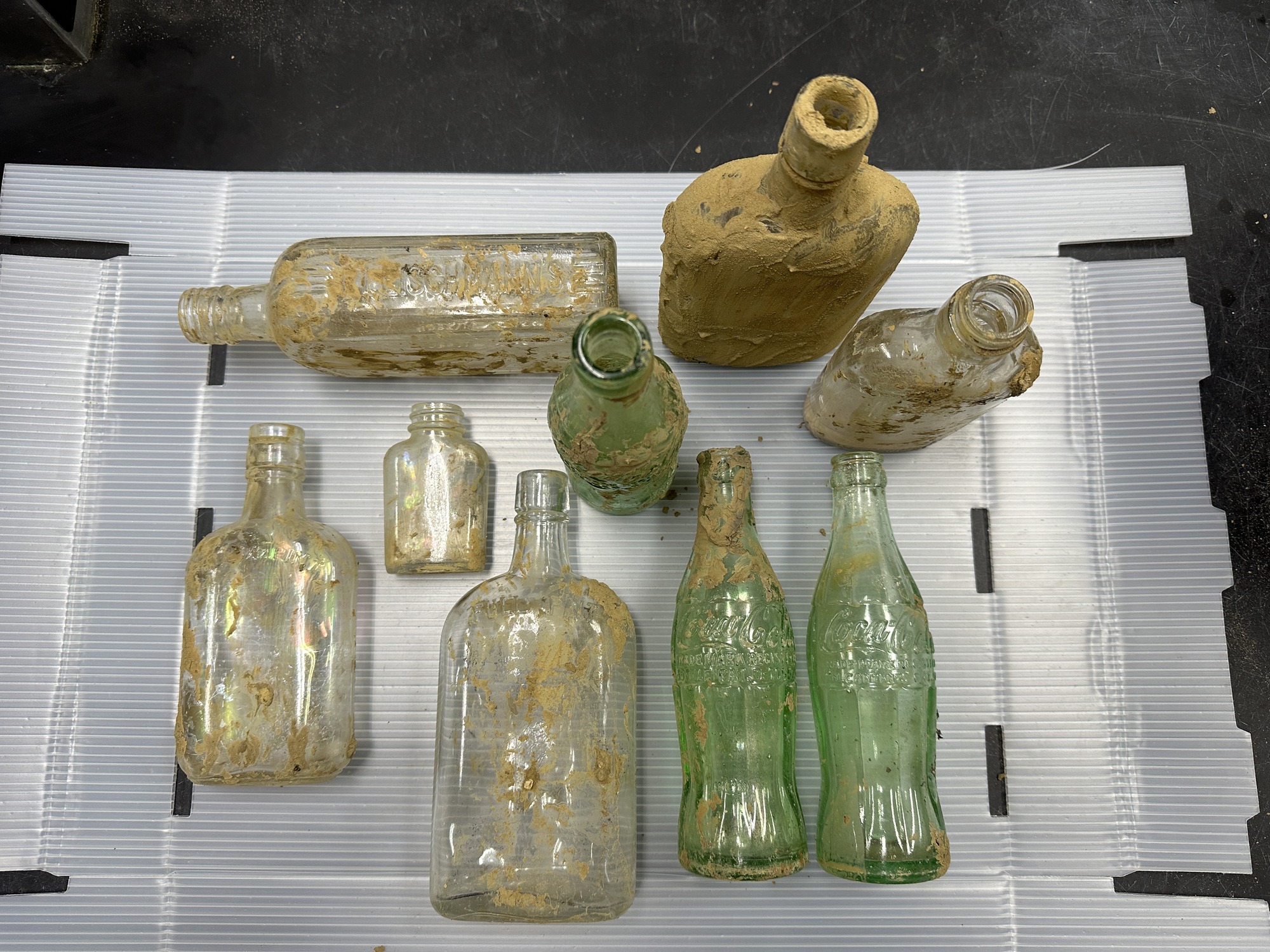Nine old glass bottles covered in mud on a display table.
