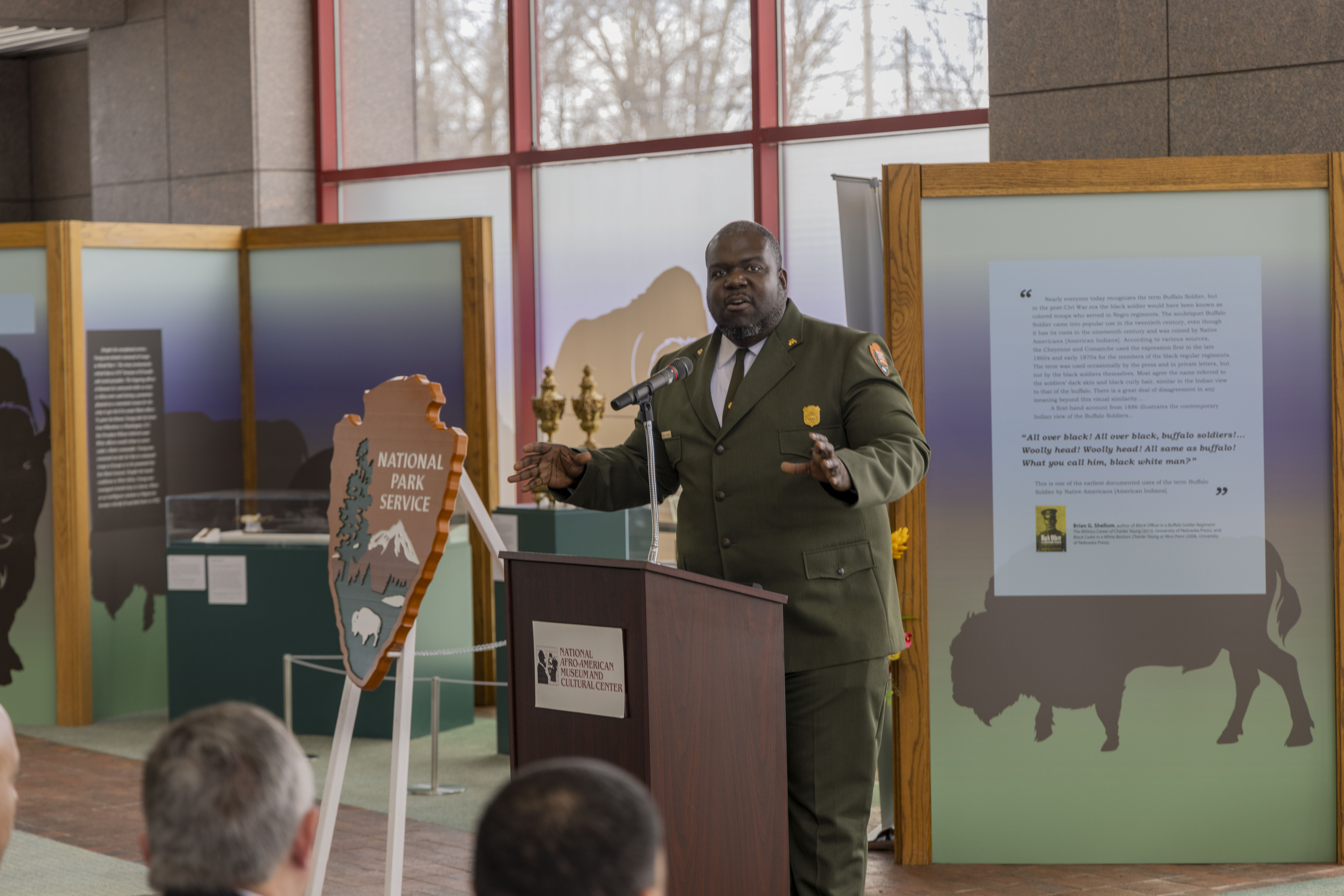A park ranger in a green suit and tie stands at a wooden podium with a microphone sticking up from it. He stands in front of a large gathering of people seated in chairs.