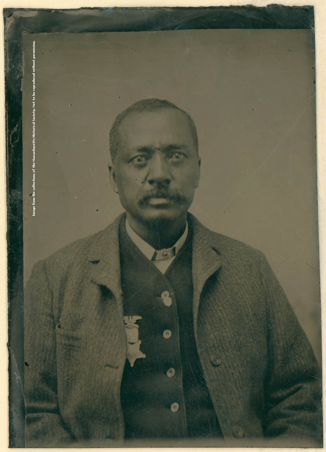 A waist up portrait of a man wearing a coat and a waistcoat, buttoned up. Shirt is a "dog collar" style. Man has a moustache and has a GAR medal pinned to his right breast on his waistcoat. The GAR medal features an eagle at the top, ribbon extending down, and a 5 pointed star hanging from ribbon.