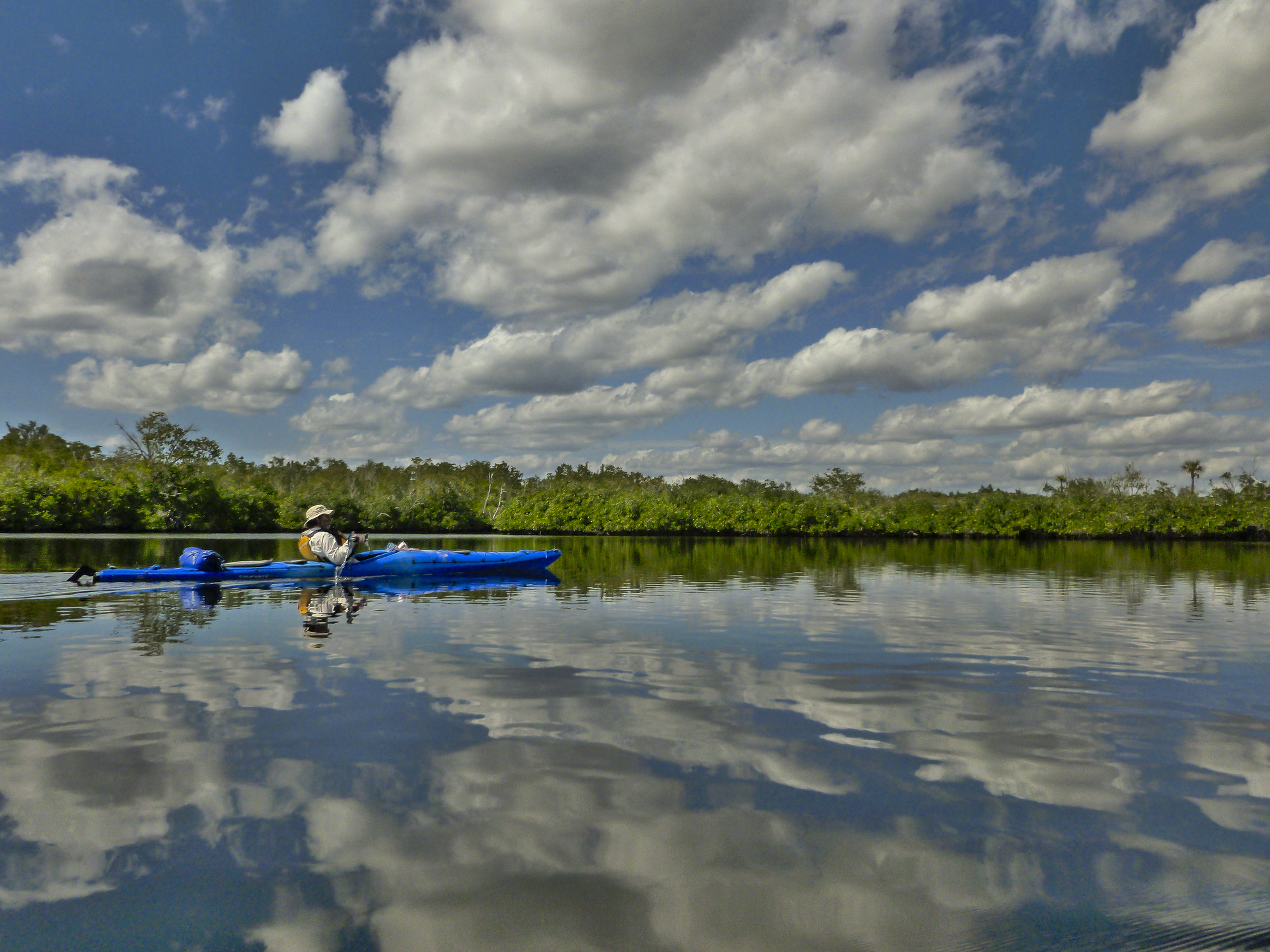 Kayaker in a wide waterway lined with trees with the clouds reflecting in the water