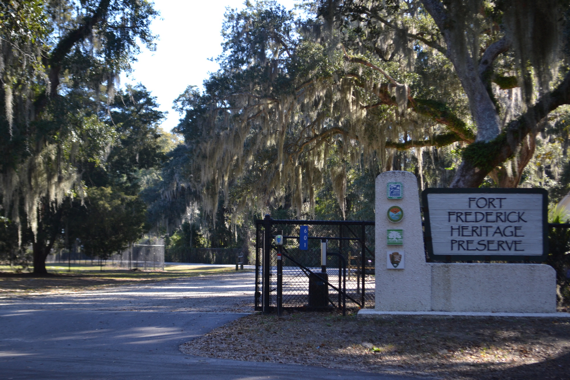 To the right of the entrance is a large wooden sign on a stone wall. The wood sign reads, “Fort Frederick Heritage Preserve.” To the left are more signs listing each of the partners. A black fence and gate sit to the left of this sign; the gate is open. The driveway is paved outside the gate. Once inside the gate, the driveway is gravel.