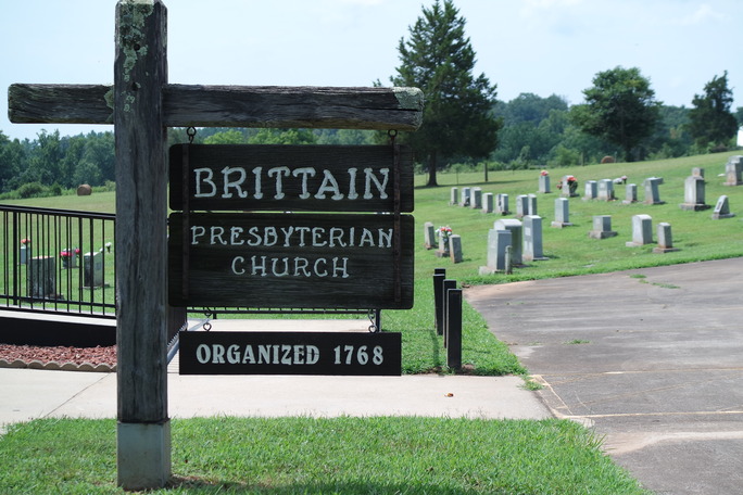 A brown wooden sign with white letters reading "Brittain Presbyterian Church, Organized 1768." In the background is a church cemetery with bright green grass and rows of grey headstones, some with bright colored flowers.