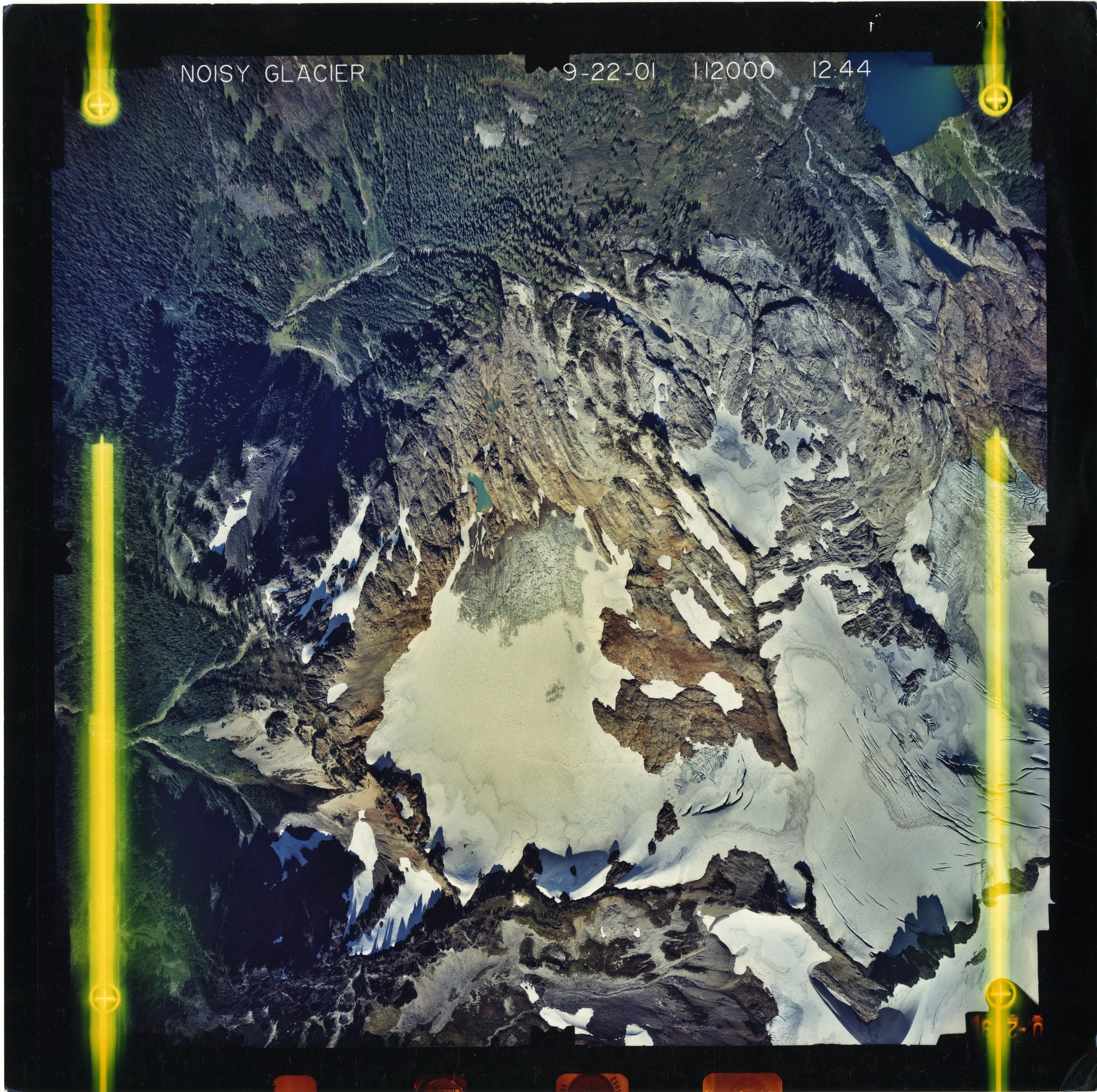 Color aerial photograph of glacier and alpine lake on mountain peak, surrounded by forested valleys.