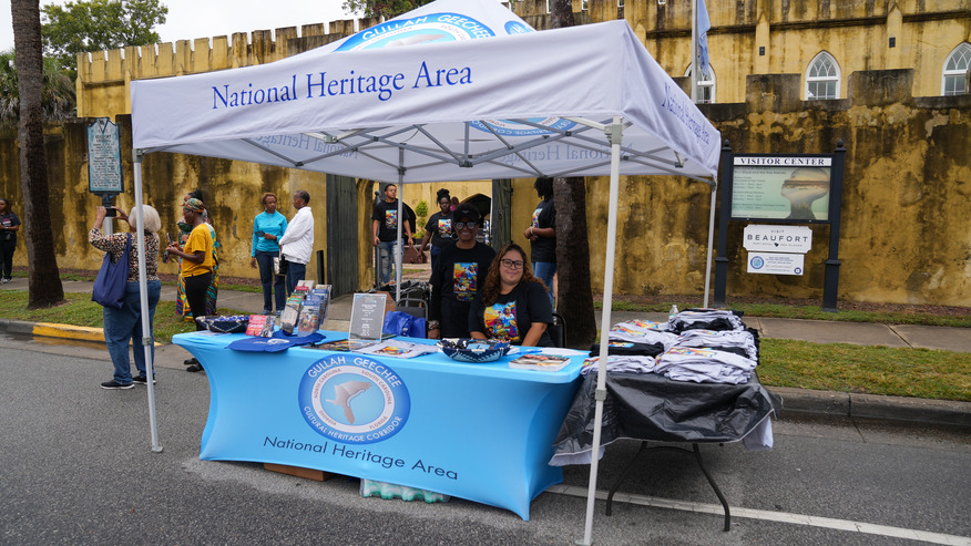 Two women sit at a tent with information table for the Gullah Geechee Cultural Heritage Corridor National Heritage Area.