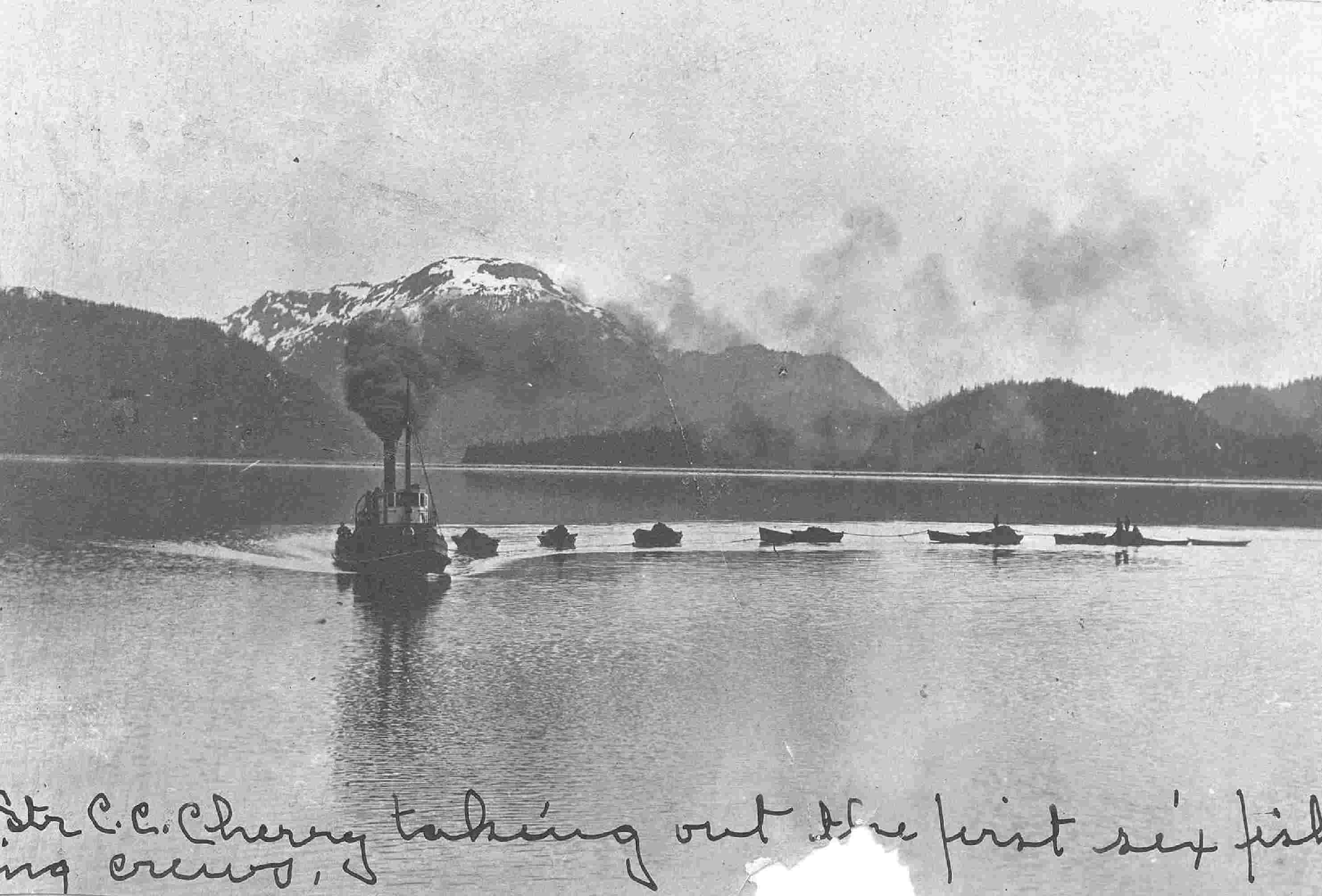 A dark puff of smoke trails behind the smokestack of a small boat, tracing the connected line of six small fishing boats silhouetted against the water. Mountains rise from the opposite shore. Black and white photograph, cursive writing on bottom says, "...C.C. Cherry taking out the first six fishing crews."