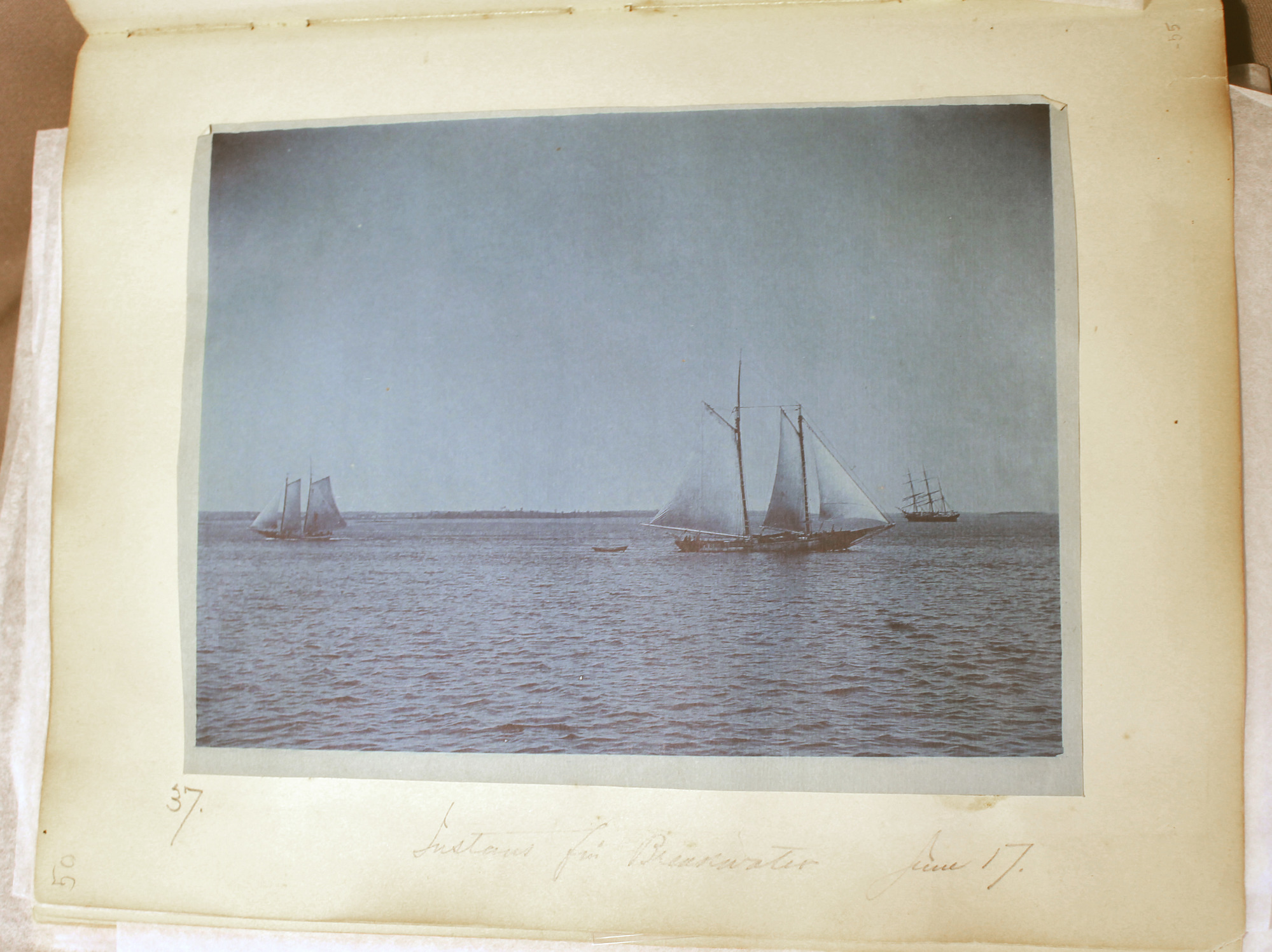 Blue toned photo of two-masted sailboat to right; two other boats with sails visible.