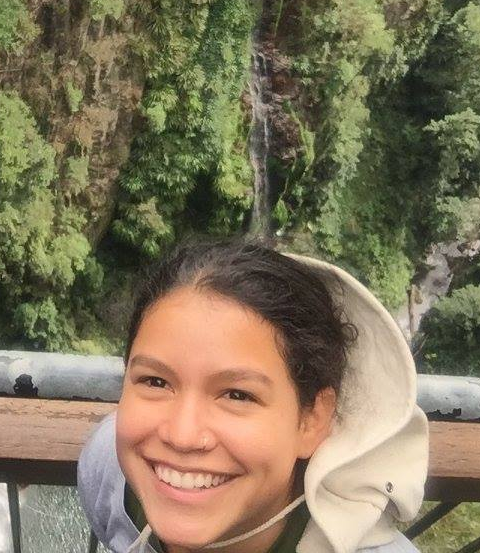 A young woman with a nose ring wearing a tan bucket hat  poses at an overlook with greenery in the background. 