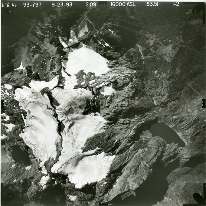 Black and white aerial photograph of glacier on mountain peak, surrounded by forested valleys.