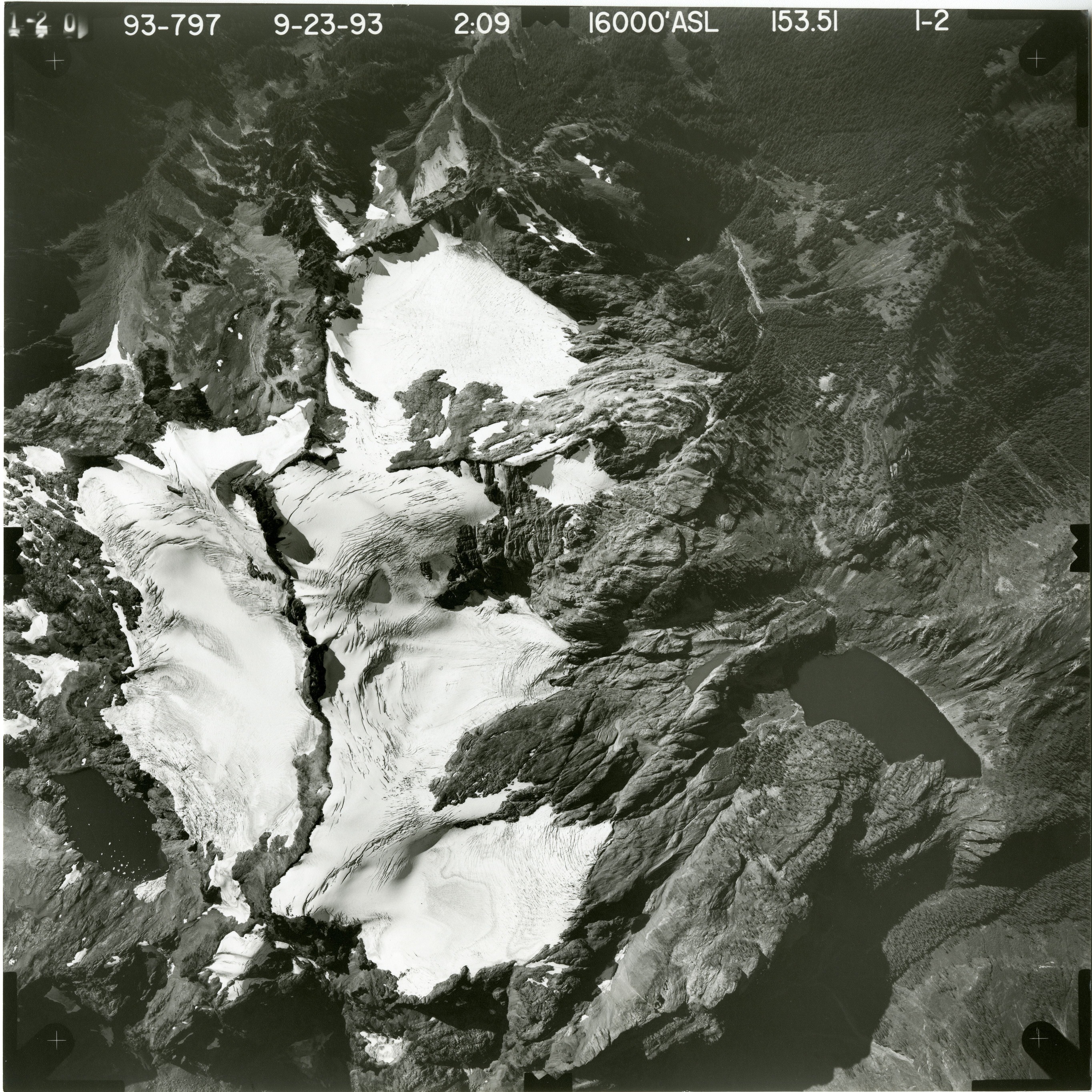 Black and white aerial photograph of glacier on mountain peak, surrounded by forested valleys.