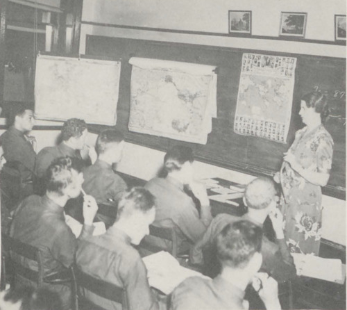 Black and white photograph of military students sitting in a classroom. Their backs are facing the camera, and all are looking at a female instructor standing at the front of the classroom. The blackboard has a variety of maps.