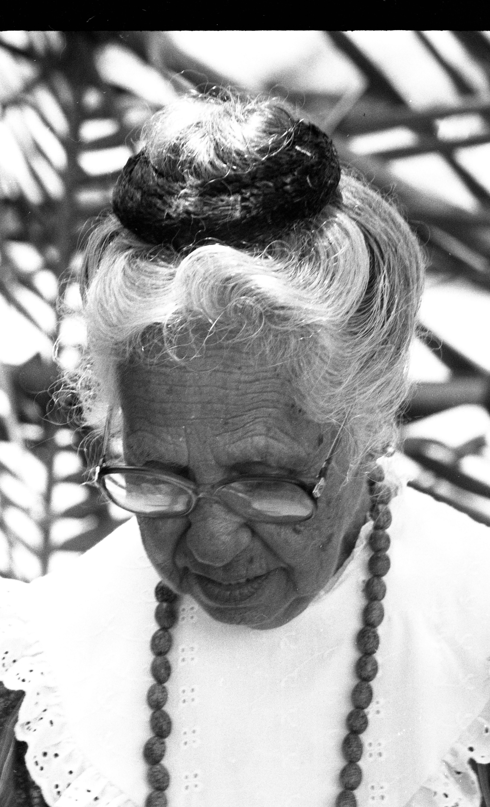 A black and white close-up head shot image of an older woman. The older woman has her gray hair up in a ballerina bun with a thick scrunchie.  She is wearing glasses and small ball-shaped earrings. She is looking downward and not at the camera. She is also wearing a beaded necklace and a high neck Vandyke eyelet collar with a scalloped hem.