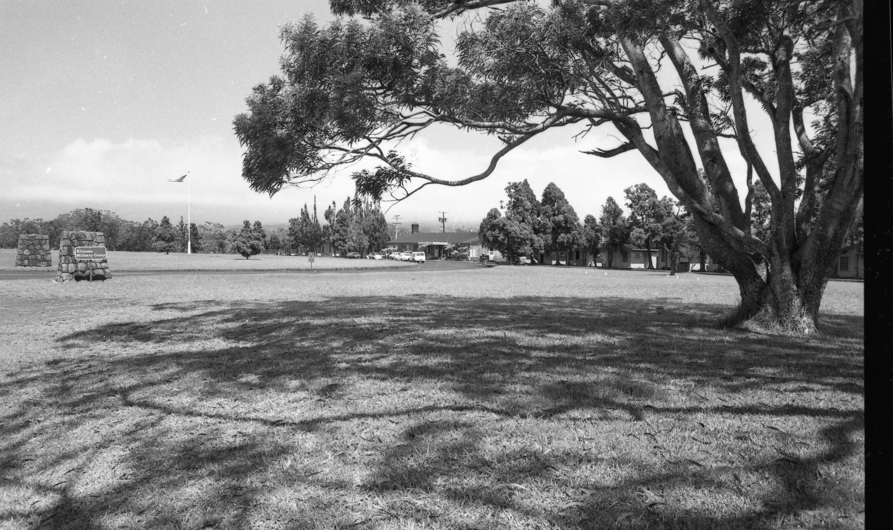 A black and white image of the front of Kilauea Military Camp from the front lawn. On the upper left side of the image there is a sign in front of a stone column that reads, "Kilauea Military Camp." Between two stone columns the entrance circle to the camp is visible. Near the sign there is a flagpole with an American flag hanging from it in the front lawn. The large front lawn in front of the cottages and main house extends from the bottom to the middle of the image. There is a large tree on the right side of the image. Towards the upper center across to the right side of the image there are cottages and the main office.  There are cars parked along a street in front of the main office and cottages.