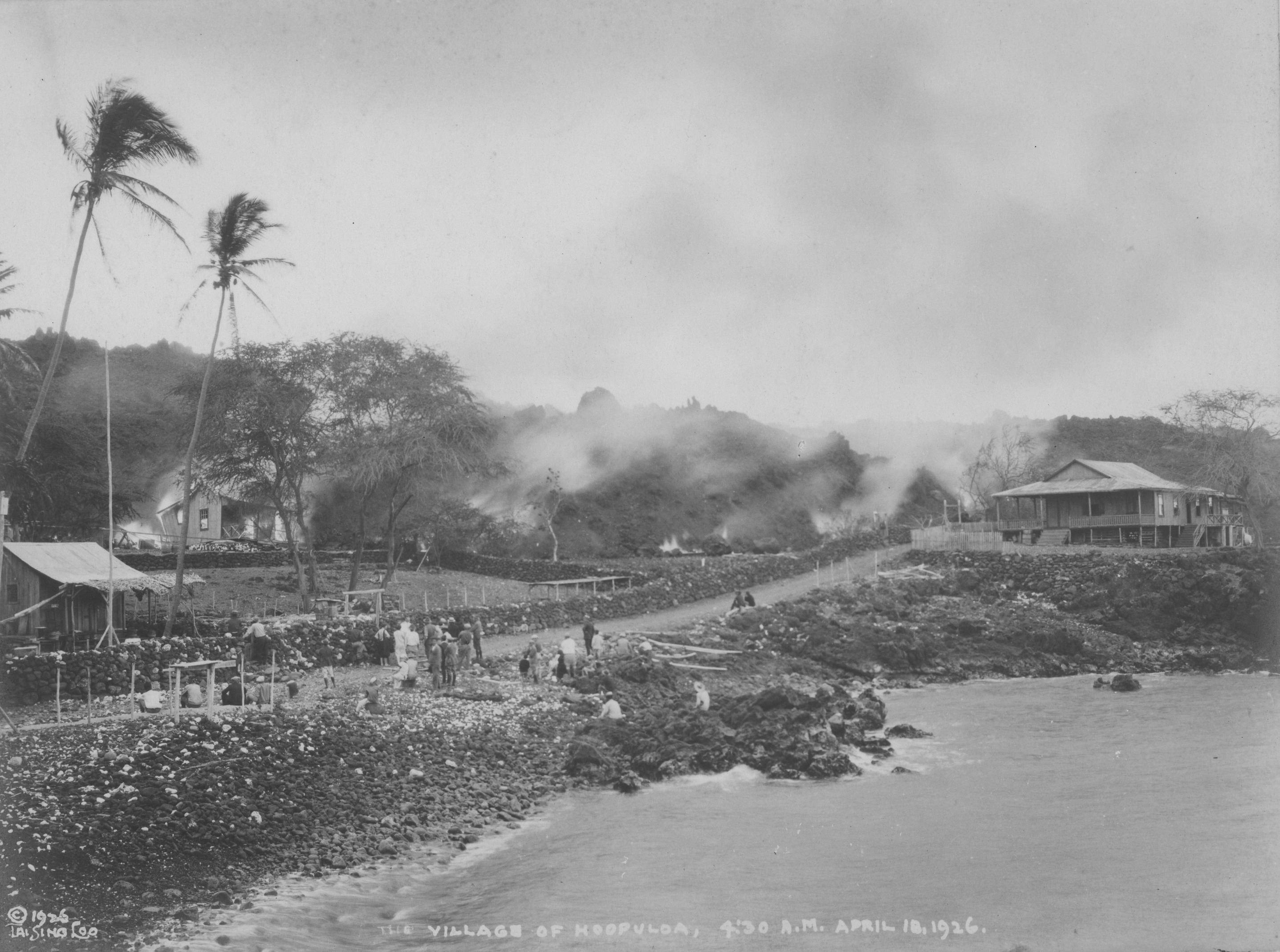 Black and white photograph of a village near the ocean. The water is present on the bottom right of the photograph near the sloped, rocky shoreline to the left. Above the water level, a dirt road runs from the left side of the image towards the right. A large house sits furthest above the water on top of a rocky hill on the right side. Many people sit and stand along the road with their backs to the camera, looking towards the elevated land to the left of the road. The elevated land emits many clouds of smoke that turn dark when rising into the sky, drifting towards the right. At the base of the elevation, on the left side, a small house is ablaze. A few tall trees are present throughout the village. Handwriting along the bottom of the photograph reads, “The Village of Hoopuloa, 4:30 A.M. April 18, 1926.” “Copyright 1926 Tai Sing Loo” is written in the bottom left corner.