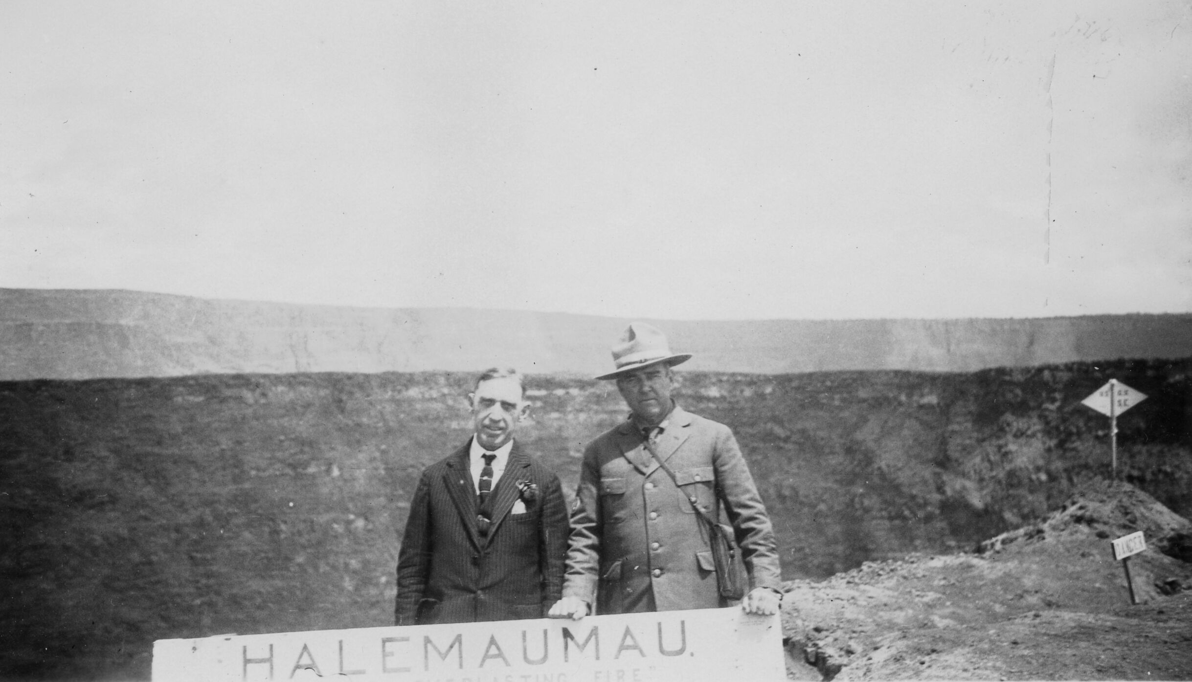 Black and white photograph of Thomas Boles (right) and an unidentified man (left) holding a sign that reads, “Halemaumau,” posing in front of the Halema’uma’u crater at Kīlauea. The man on the left is dressed in formal attire, and Boles wears National Park Service attire. The two men stand in the middle of the photograph at the forefront and the sign is only partially visible, where the words “Halemaumau” and “Fire” can be seen. Behind the two men is a large crater that extends the entire middle of the photograph with elevated land in the background. Towards the right side of the image, rough, rocky terrain is present along with two signs on wooden posts. A rectangular sign closest to the camera reads “DANGER” and a diamond-shaped sign behind it is illegible.