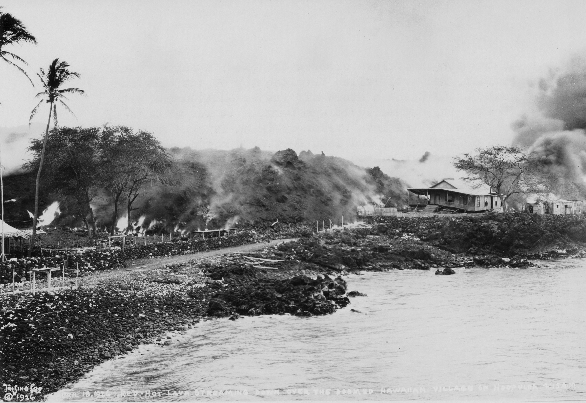Black and white photograph of a village near the ocean. The water is present on the bottom right of the photograph near the sloped, rocky shoreline to the left. Above the water level, a dirt road runs from the left side of the image towards the right. A large house that sits furthest above the water on top of a rocky hill towards the right side is being damaged by a lava flow. Elevated land to the left emits clouds of smoke and areas of fire, reaching the road. Behind the house to the left, a large, dark cloud of smoke rises. There are still spots of standing vegetation before the lava eventually reaches the water. Handwriting along the bottom reads, “April 19, 1926, Red-Hot Lava Streaming Down Over The Doomed Hawaiian Village Of Hoopuloa 4:15A.M.” “Tai Sing Loo, copyright 1926” is written in the bottom left corner.