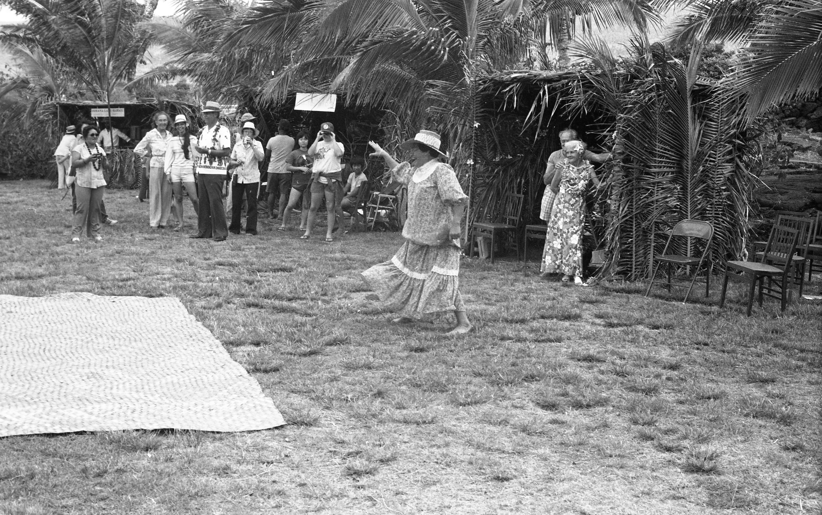 A black and white image of a dancer performing at an event. She is wearing a hat and a short sleeve dress with a ruffled collar. Her legs are far apart, her right arm is raised out to the side while her left arm rests at her side. She is facing the left side of the image. There is a woven mat on the grass in front of her. On the far-right side of the image there are three chairs, one metal and two wooden. To the left of the chairs is a shelter covered in palm leaves. There is a man and woman standing just inside the shade shelter. The woman has a flower tucked behind her left ear. She is wearing a short sleeve dress, sandals, and a lei around her neck. The man is standing behind her wearing a t-shirt and shorts. He is holding onto her shoulder with his right hand. There are two wooden chairs to the left of them. There are two other shade shelters next to this one extending to the left side of the image. These shelters have signs hanging from the roof. Six people are standing in front of the shelters watching 