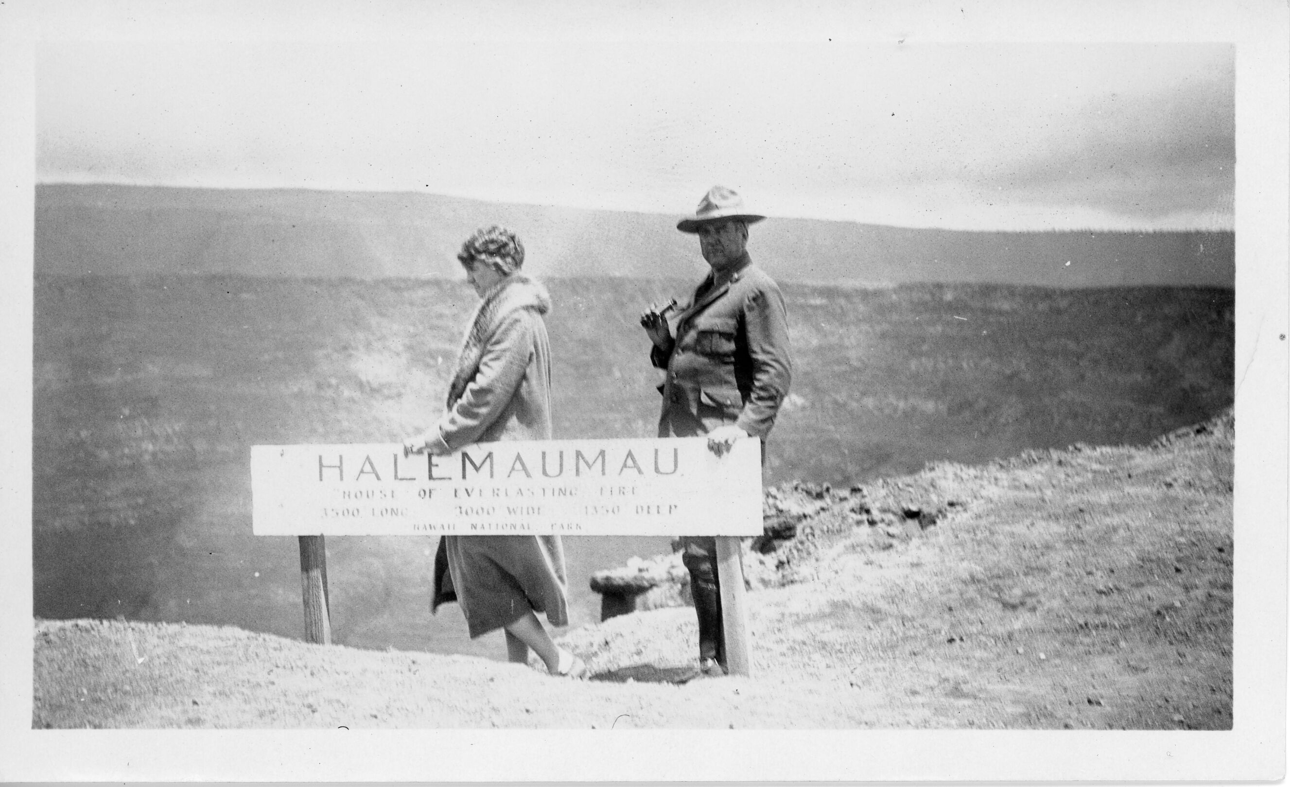 A black and white image of Inez and Thomas Boles standing behind a sign for Halemaʻumaʻu. The Boles are pictured in the center of the image standing behind a wooden sign. The sign reads, “Halemaʻumaʻu, House of Everlasting Fire, 3500' long, 3000’ wide, 1350’ deep, Hawaii National Park.” Inez is located behind the center of the sign. She is wearing a long fur lined coat with her hair up. She is holding onto the sign with her left hand and is facing the left side of the image. Thomas is standing behind her also holding onto the sign with his left hand. He is facing the left side of the image with his head turned facing the camera. He is wearing a hat and a suit. He is holding a pair of glasses in his right hand. In the background, the walls of the crater are visible.