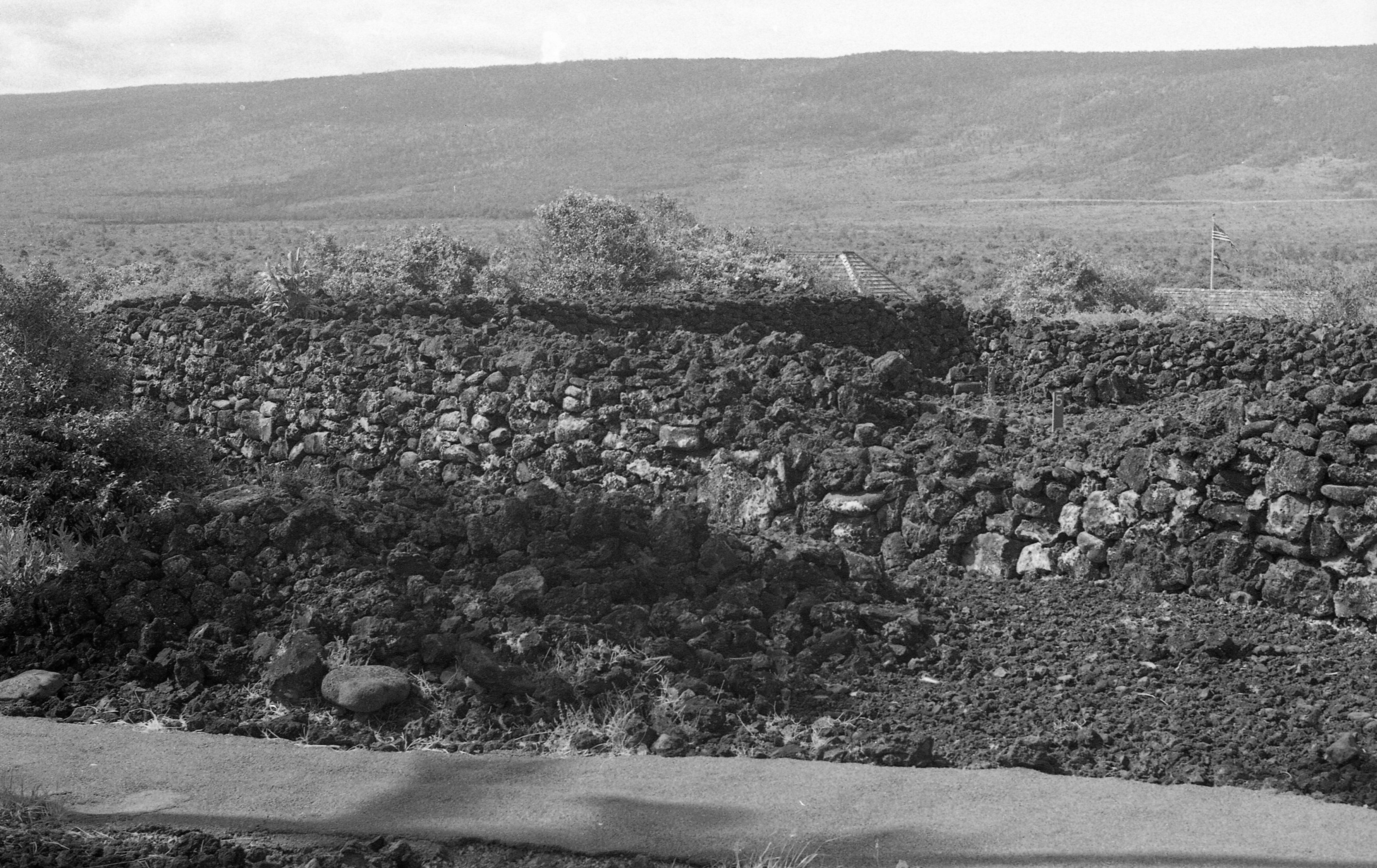 A black and white image of lava rock walls. There is a path on the bottom of the image that extends from the left side to the right side of the image. The lava rock walls are located behind the path.  Each of the three lava walls connect to form a square shape. Towards the center right side of the image there is a wooden post with the number "5" painted onto it. The post is in the middle of the three walls.  In the distance towards the top right corner of the image is an American flag hanging on a flagpole. There are also roofs of two buildings immediately behind the farthest wall. In the background there is a mountain.