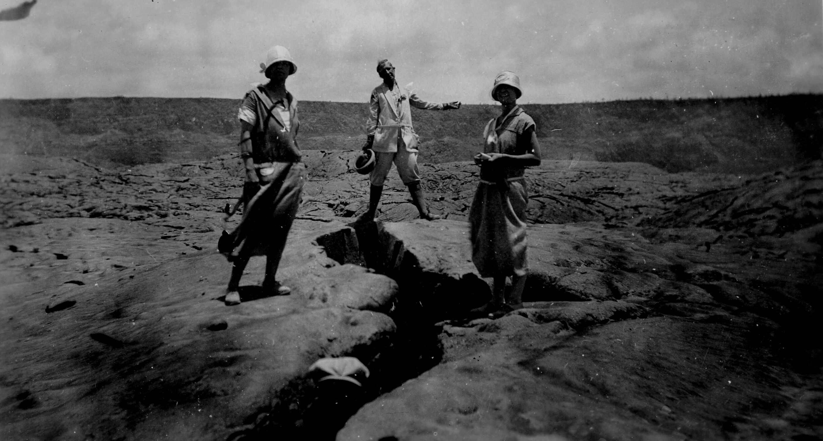 Black and white photograph of two women and one man posing for the camera, standing around a large crack within lava bedrock. The photograph is dark, and it appears there may be another person standing inside of the crack close to the camera, as evidenced by an outline of a hat. Both women are standing to the left and right of the crack, and the man is standing in the center of the image straddling the crack with both legs and an outstretched arm. The terrain is dry and barren.
