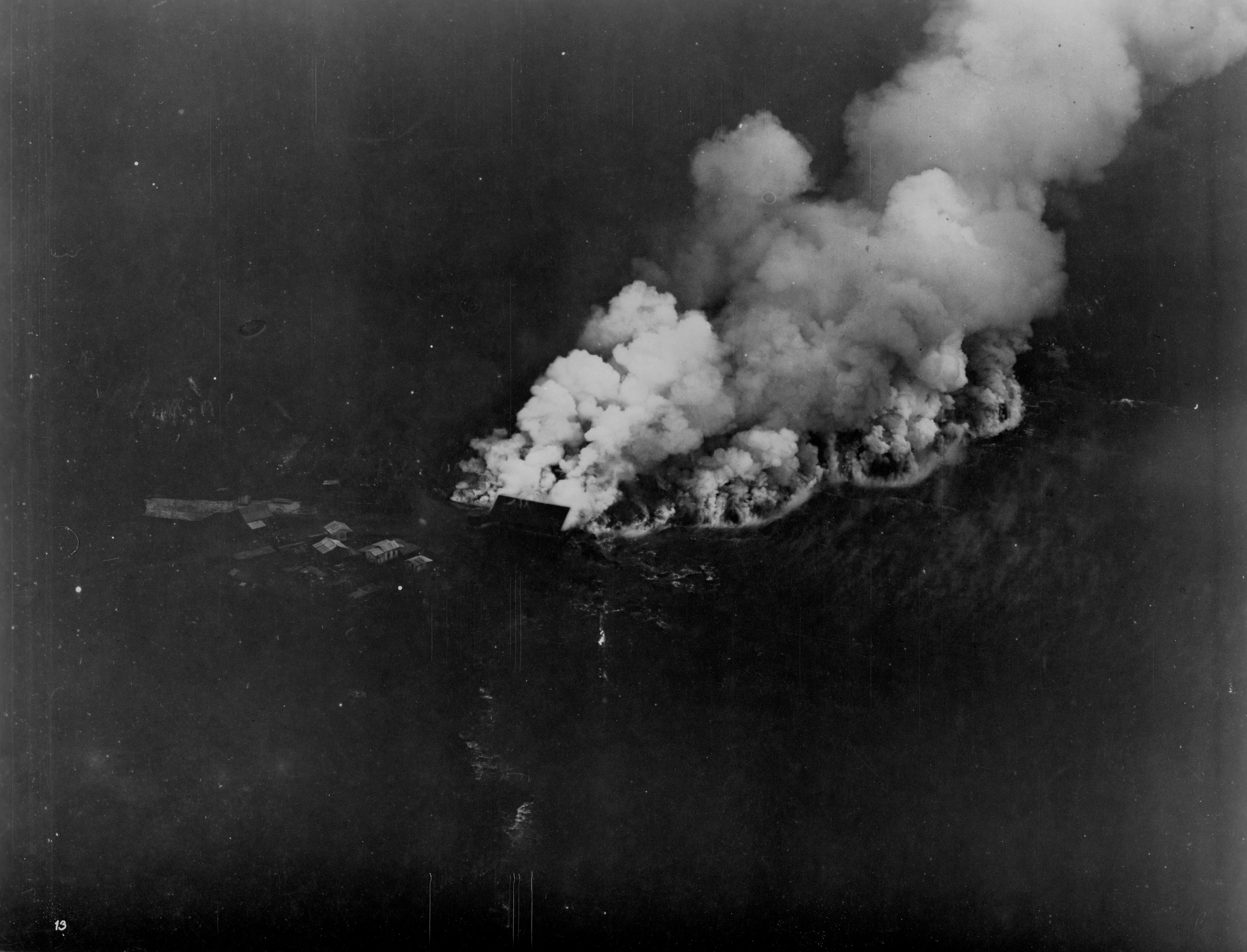 Black and white photograph of large plumes of smoke taken from a bird’s-eye view. The photograph is dark, and only the smoke and some buildings are visible. The white smoke cloud begins at the center of the photograph and extends beyond the top right corner. To the left of this, A small group of buildings can be seen on the ground. The rest of the photograph is almost completely black. A handwritten number “13” is present in the bottom left corner.