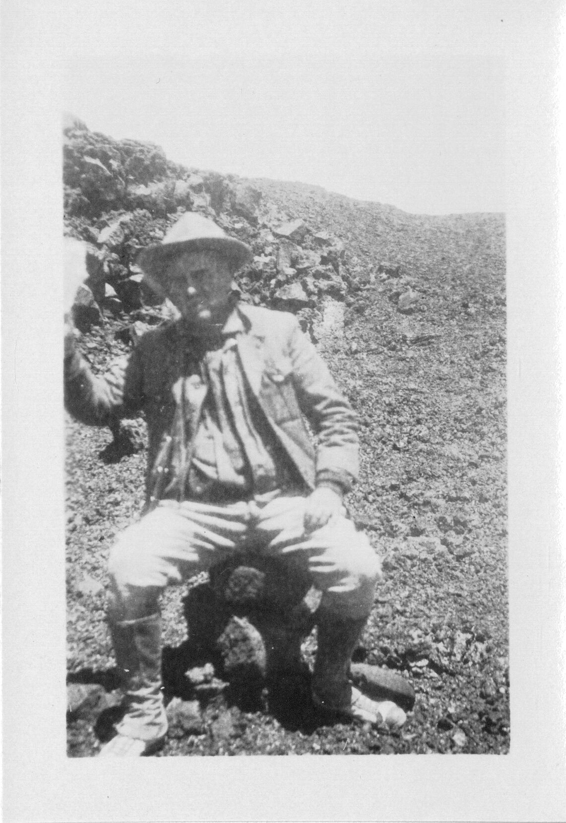 A black and white image of Thomas Boles resting on the hike to Mauna Loa’s summit. Boles is pictured in the center of the image. He is wearing a hat, jacket, button down shirt, and jeans. He is sitting on a stack of rocks and is looking towards the camera. His left hand is resting in his lap while his right hand is raised either waving or holding an unidentified object out of frame. In the background there is a slope covered in ʻaʻā lava.